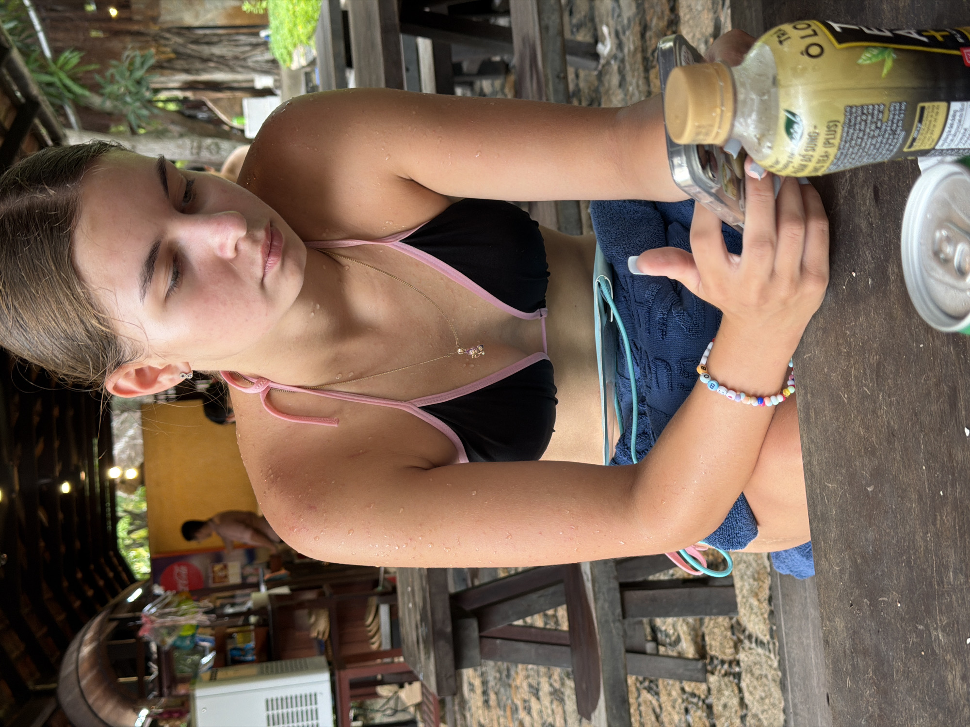 A young Caucasian woman with brown hair pulled back is sitting at a rustic wooden picnic table, looking down at her mobile phone. She is wearing a black bikini with pink trim and a gold necklace with a small pink charm. Her skin is wet, suggesting she has been in the water. She has a beaded bracelet on her left wrist. A blue towel is draped over her lap. To her right, a bottle of "O Long Tea Plus" is on the table, next to a silver can. In the background, wooden picnic tables and benches are visible. The scene appears to be outdoors under a thatched roof, possibly at a beachside cafe or resort in Nha Trang, Vietnam, given the lush greenery and tropical trees visible in the distance. A white refrigerator unit is to the left. Further back, a man in a light-colored shirt is standing near a yellow wall. The ground appears to be gravelly.