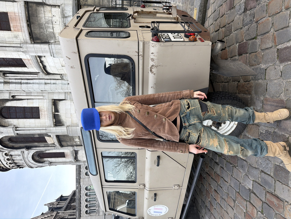 A blonde female, appearing to be a young adult, stands centrally on a cobblestone surface, facing the camera with a neutral expression. She wears a bright blue flat-topped cap, a brown corduroy jacket with an asymmetric button closure, a black crop top, heavily distressed light blue jeans with dark patches, and light tan boots. A black strap crosses her chest, and she holds a lit cigarette between the thumb and index finger of her left hand.
Directly behind her is a light khaki Land Rover Defender 110. A circular sticker displaying "VIGIPIRATE" and a tricolor emblem is affixed to its driver's side door. The rear of the vehicle shows a partially visible license plate displaying "G009 0049" and red taillights. A reflection of a dark-haired individual wearing a dark top is visible in the vehicle's rear windows.
In the background, the distinctive white stone facade of the Basilica of the Sacré-Cœur, Montmartre, Paris, France, with its prominent domes, arches, and windows, is visible under an overcast sky. A smaller, darker stone building with similar architectural elements is visible to the left of the main basilica. The ground is paved with cobblestones.