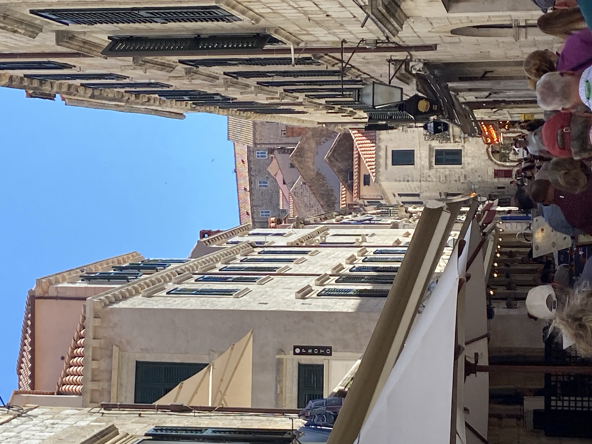 This is a photograph taken on a sunny day in Dubrovnik, Croatia, likely within the Old Town. The image captures a narrow street lined with ancient stone buildings featuring characteristic terracotta roofs and dark-shuttered windows.
On the left side of the street, a building displays a sign that reads "PROTO" vertically. This building has awnings extended over what appears to be an outdoor seating area, possibly a cafe or restaurant. Several white umbrellas are visible, providing shade to tables below. Some clothing items are also hanging outside this building.
On the right side of the street, another building exhibits a more traditional facade with multiple windows and decorative architectural elements. A vintage-style street lamp with a dark metal frame and a glass enclosure hangs from this building. Further down the street, the architecture becomes more varied, with buildings of different heights and roof styles visible, suggesting the complex urban fabric of the city.
The street is populated by a number of people walking, some are clustered in groups, suggesting a tourist area. They are dressed in casual summer clothing. The sky is a clear, bright blue, indicating fair weather. The overall scene conveys a sense of a bustling, historic European city.