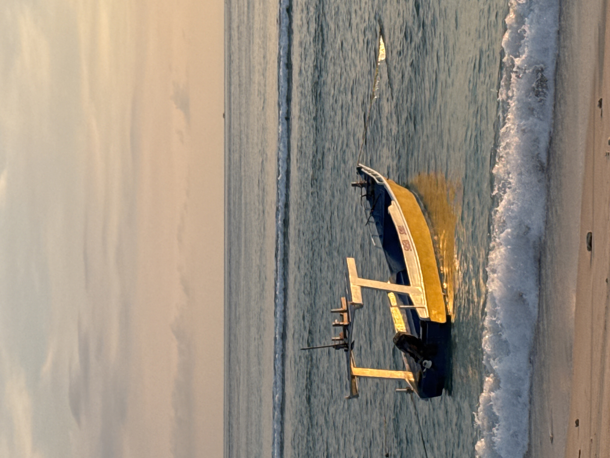 The image shows a yellow and blue outrigger boat docked on a sandy beach in Unknown, Indonesia. The boat is partially submerged in the water, near the shoreline where a wave is breaking. The sky is overcast, with a light, diffused glow. The sea is calm, with gentle waves visible further out.