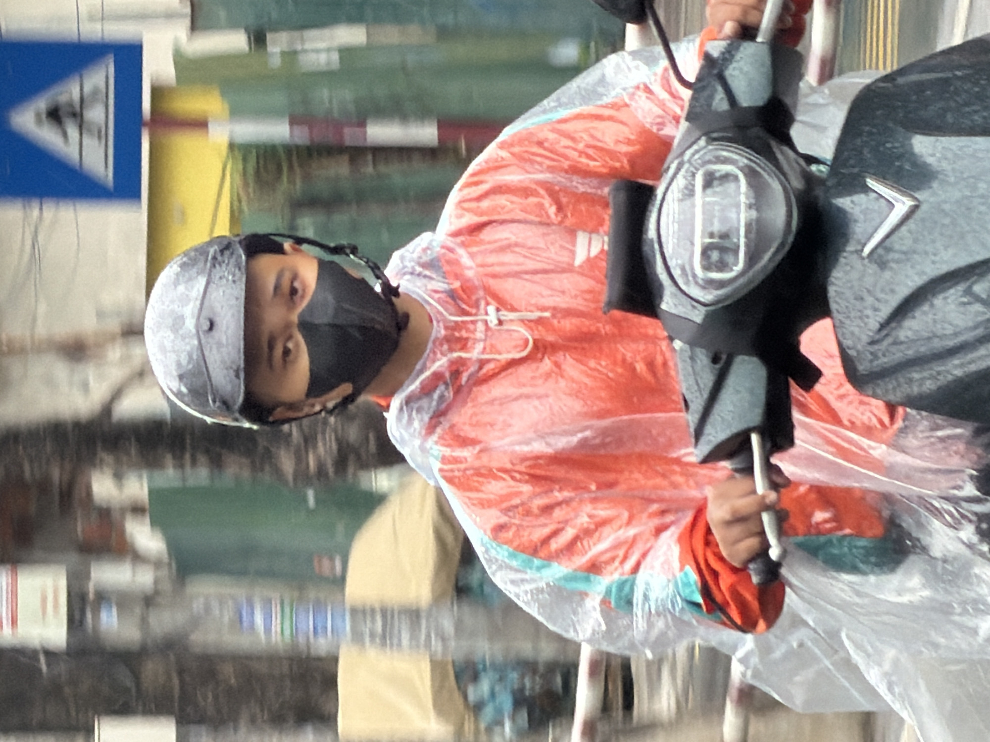 A person wearing a black mask and a gray helmet rides a dark-colored scooter. The person is wearing a bright orange jacket over a clear poncho. The person's hands are on the scooter's handlebars. In the background, there are blurred buildings, trees, and a blue and white road sign. The scooter has a V-shaped emblem on its front. The image appears to be taken on a street in Nha Trang, Vietnam, as indicated by the presence of a road sign common in Vietnamese cities. The overall scene depicts a person commuting on a scooter in an urban environment, possibly during rainy weather due to the poncho.
