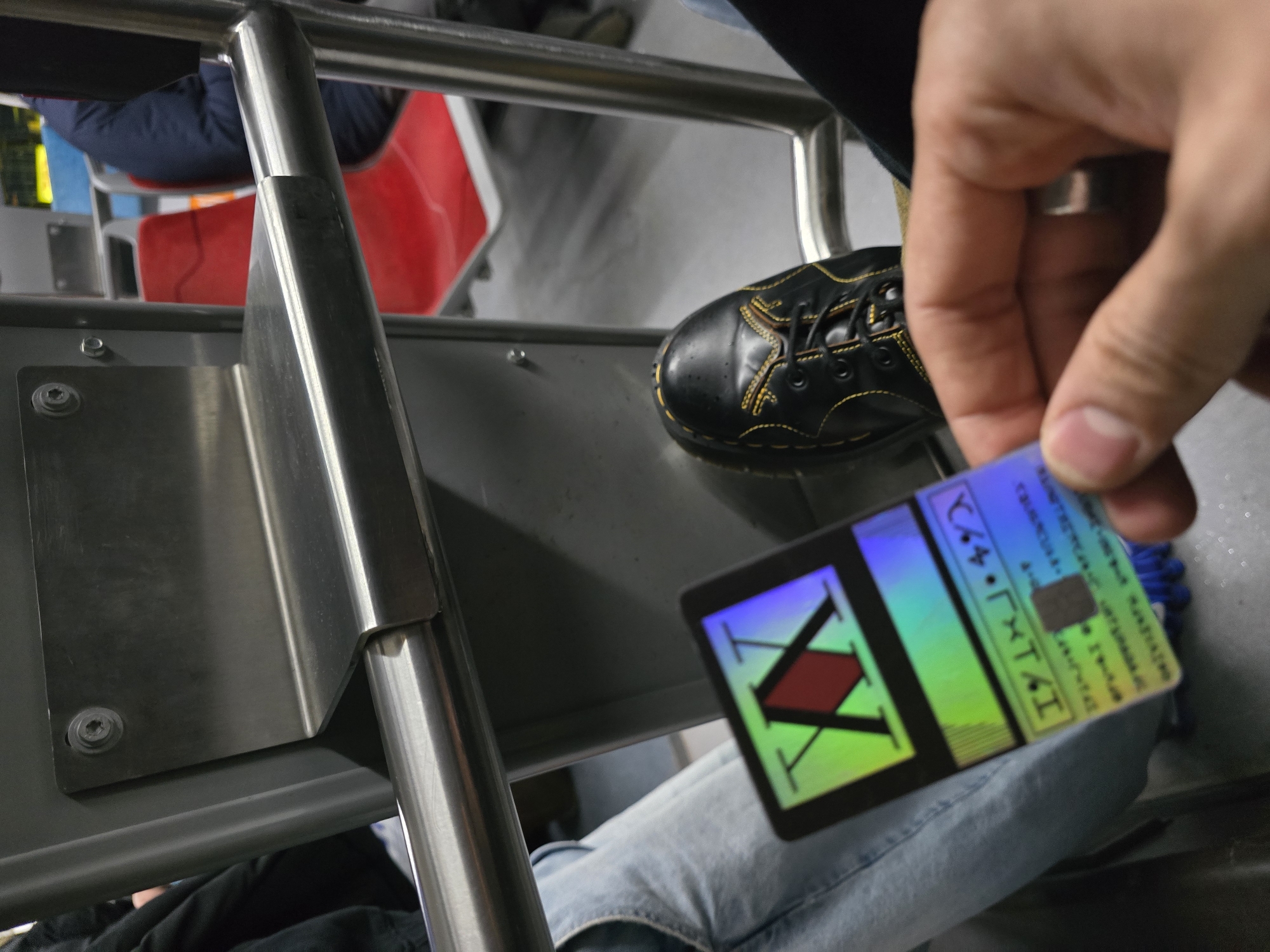 A person is on a train in Aubervilliers, France, holding a Hunter x Hunter hunter license card. The person is wearing light blue jeans and black Dr. Martens shoes. There is a silver ring on the person's finger. A red seat is visible in the background. The train car has silver metal bars and grey seats.