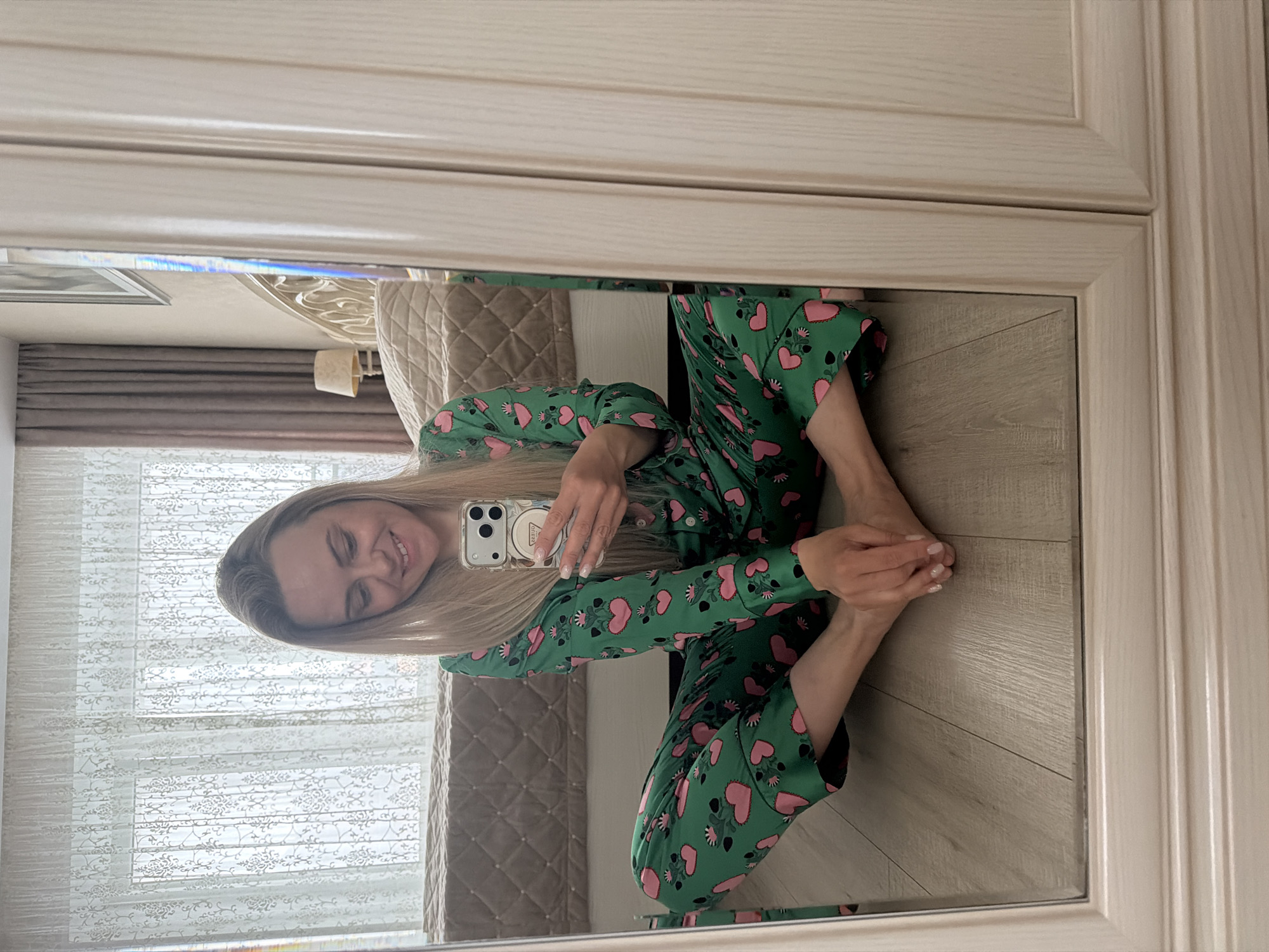 This image, taken in Votkinsk, Russia, shows a woman taking a mirror selfie. The woman, with long blonde hair, is seated on the floor, captured in the reflection of a large, light-framed mirror. She is smiling broadly, with her eyes slightly closed, conveying a sense of happiness or relaxation.
She is dressed in a vibrant green pajama set patterned with numerous pink hearts and subtle floral elements. Her legs are crossed, and she holds a smartphone, likely an iPhone given its camera configuration, in front of her. The phone has a clear case with a small white circular charm or sticker on its back. Her fingernails are manicured with a light, possibly glittery, polish.
The background, visible as a reflection in the mirror, shows parts of a room. To the left, a window covered by white sheer curtains with a floral pattern allows natural light into the space, suggesting it is daytime. A beige quilted bed headboard and a small lamp with a light-colored shade are also reflected. The floor beneath her in the reflection is light-colored wood. The mirror itself is encased in a light, possibly wooden, frame that appears to be part of a wardrobe or interior door. No distinct legible text is visible in the image.