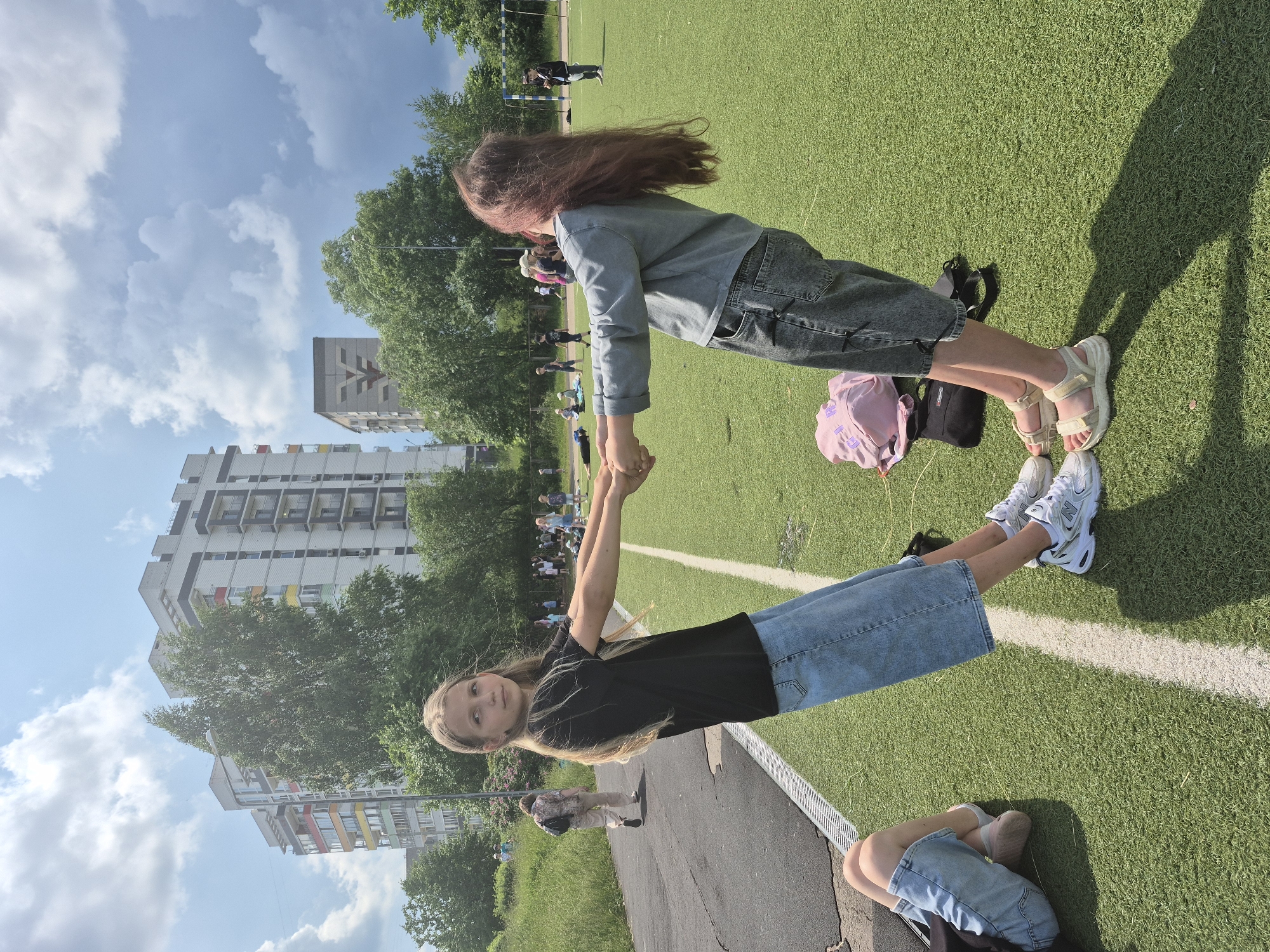 Two young girls are holding hands and leaning backwards on a bright green artificial turf field, which appears to be a sports field or playground in Chaikovsky, Russia. The girl on the left has long blonde hair and is wearing a black t-shirt and a light blue denim skirt. The girl on the right has long, dark reddish-brown hair and is wearing a grey long-sleeved shirt and dark grey denim shorts. Their bodies are angled away from each other, creating a V-shape between them. Their feet are positioned widely apart for balance.
In the foreground, a third person's legs are visible, lying down on the grass to the left. This person is wearing light blue denim shorts or capris and a dark top. They appear to be on their side, with their legs bent.
In the background, there are several multi-story apartment buildings with white facades and colorful accents. Trees and greenery are scattered throughout the scene. Other children and adults are visible in the distance, some walking and some gathered in groups, suggesting a public area or event. A football goal is also visible on the right side of the image. The sky is blue with scattered white clouds, indicating a sunny day. A pink backpack and a black bag are on the grass near the girl on the right.