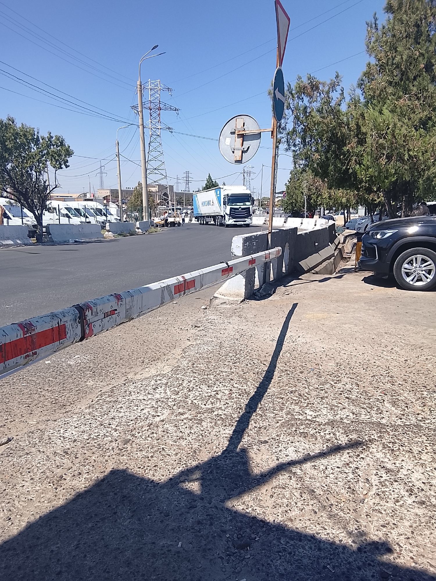 The image captures a street scene in Tashkent, Uzbekistan, under a clear blue sky. In the foreground, a concrete barrier with red and white striped markings separates the road from a raised area. The road itself is paved and appears to be a thoroughfare.
Further down the road, several white vans are parked in a row, suggesting a commercial or industrial area. A large white truck with a blue and white logo on its side, bearing the text "HOWO-MAX," is moving away from the viewer.
To the right of the road, a dark-colored SUV is parked. Behind the concrete barrier, trees line the side of the road, casting shadows. In the background, a prominent power pylon with overhead cables indicates the presence of significant electrical infrastructure. Traffic signs, including a triangular sign with a red border and a circular sign with an arrow, are visible on a pole near the trees. The overall environment suggests a relatively busy urban setting.