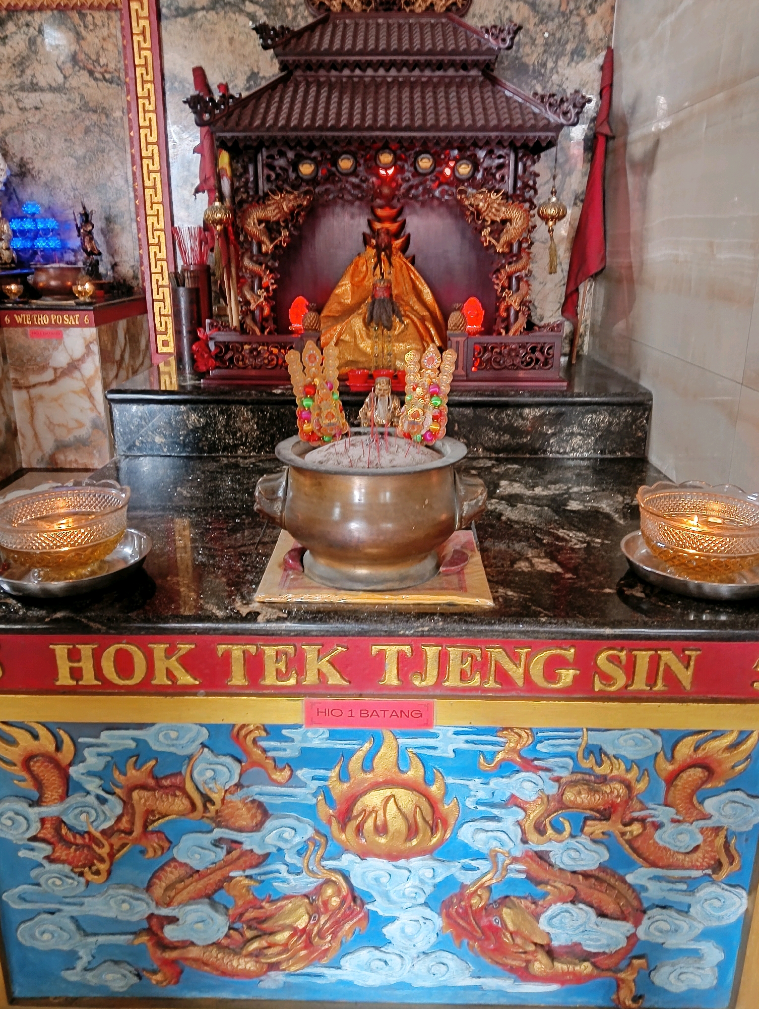 The image depicts an altar, likely in a temple or shrine, featuring religious iconography. At the center of the altar is a golden statue of a deity, draped in flowing robes. Incense sticks are burning in a bronze censer placed in front of the statue. Flanking the censer are two ornate bowls, one on each side, containing lit candles. The altar itself is adorned with carvings of dragons and other mythical creatures, and is surrounded by decorative elements. A red banner with gold lettering is visible below the altar. The background consists of marbled walls and a wooden structure, typical of religious spaces. The scene suggests a place of worship or offering.
Based on the provided text "HIO 1 BATANG" and "HOK TEK TJENG SIN" visible on the altar, this location is likely a Hok Tek Tjeng Sin temple. Temples dedicated to Hok Tek Tjeng Sin, a deity associated with prosperity and fortune, are common in Chinese communities in Indonesia. Without further information to pinpoint the exact location, it is impossible to confirm its presence in the city of Rangkasbitung, Indonesia.