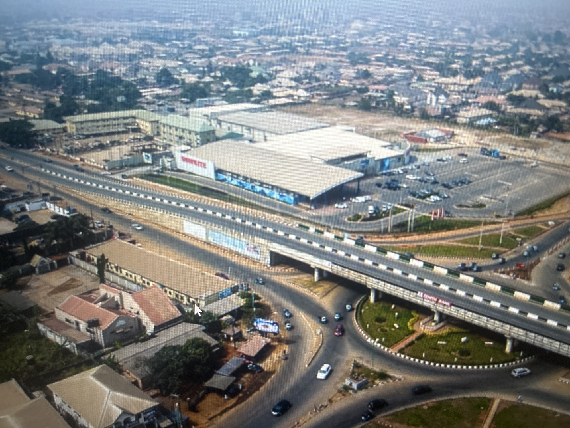This is an aerial view of the Shoprite supermarket and surrounding area in Ogbomosho, Nigeria. A large, modern Shoprite store with a prominent red and white sign is visible, along with its extensive parking lot filled with cars. Adjacent to the supermarket is a multi-lane road with a pedestrian bridge crossing over it. Several buildings, including residential homes and what appear to be other commercial establishments, are scattered throughout the urban landscape. Cars are visible on the roads and in the parking lot, indicating active traffic and economic activity. The scene depicts a typical urban environment with commercial infrastructure, transportation networks, and residential areas.