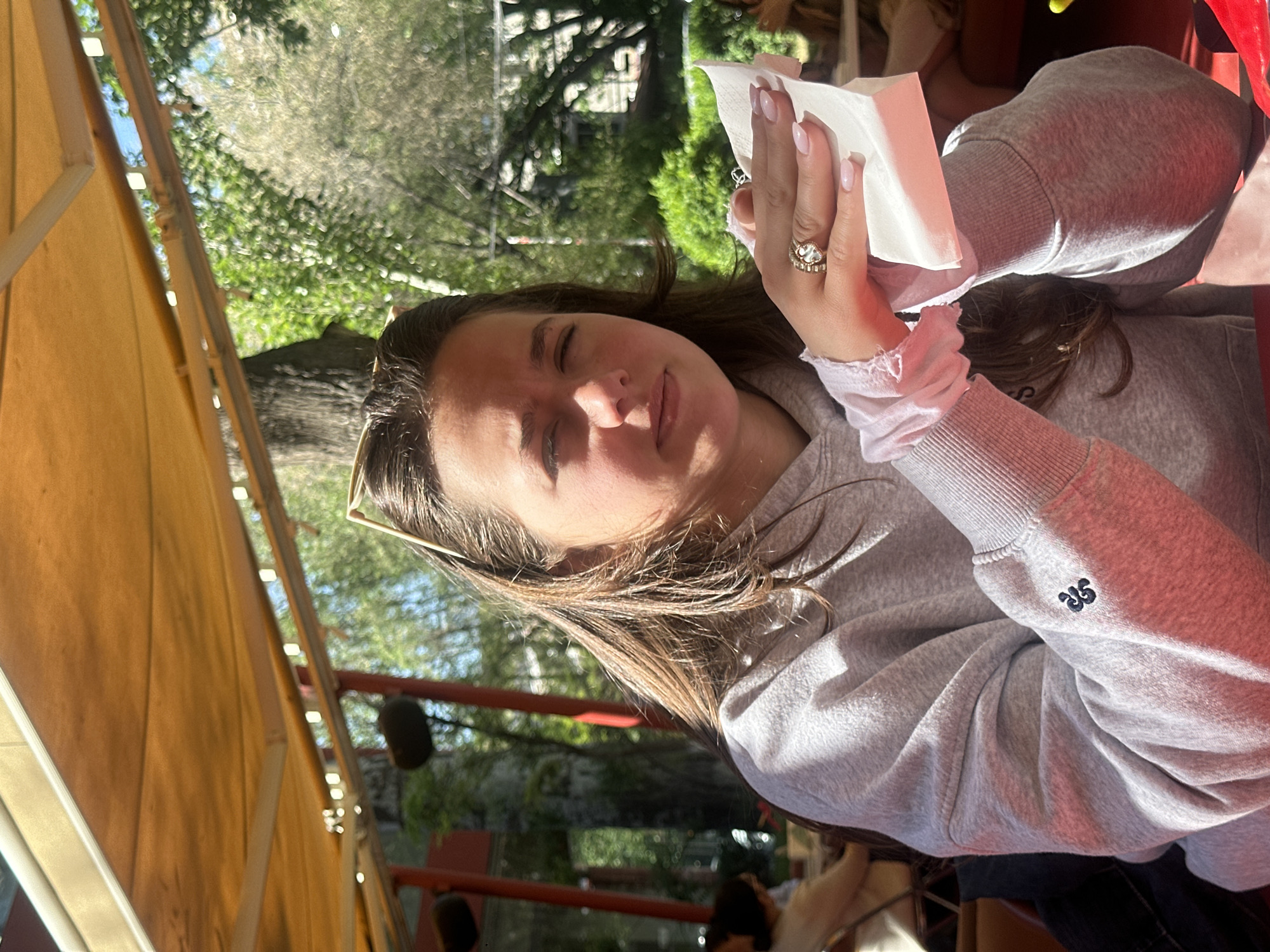 A young woman is pictured in an outdoor setting. She is holding a white tissue in her right hand, adorned with a large, ornate ring. Her left hand is partly visible, also bearing a ring. She is wearing a grey sweatshirt with an embroidered design on the sleeve. She seems to be seated, with a glimpse of a table and potentially other people in the background. The background shows greenery, suggesting a park or garden environment. The presence of a yellow awning or structure indicates the setting might be a cafe or restaurant terrace. There is no verifiable indication of Yerevan, Armenia.
