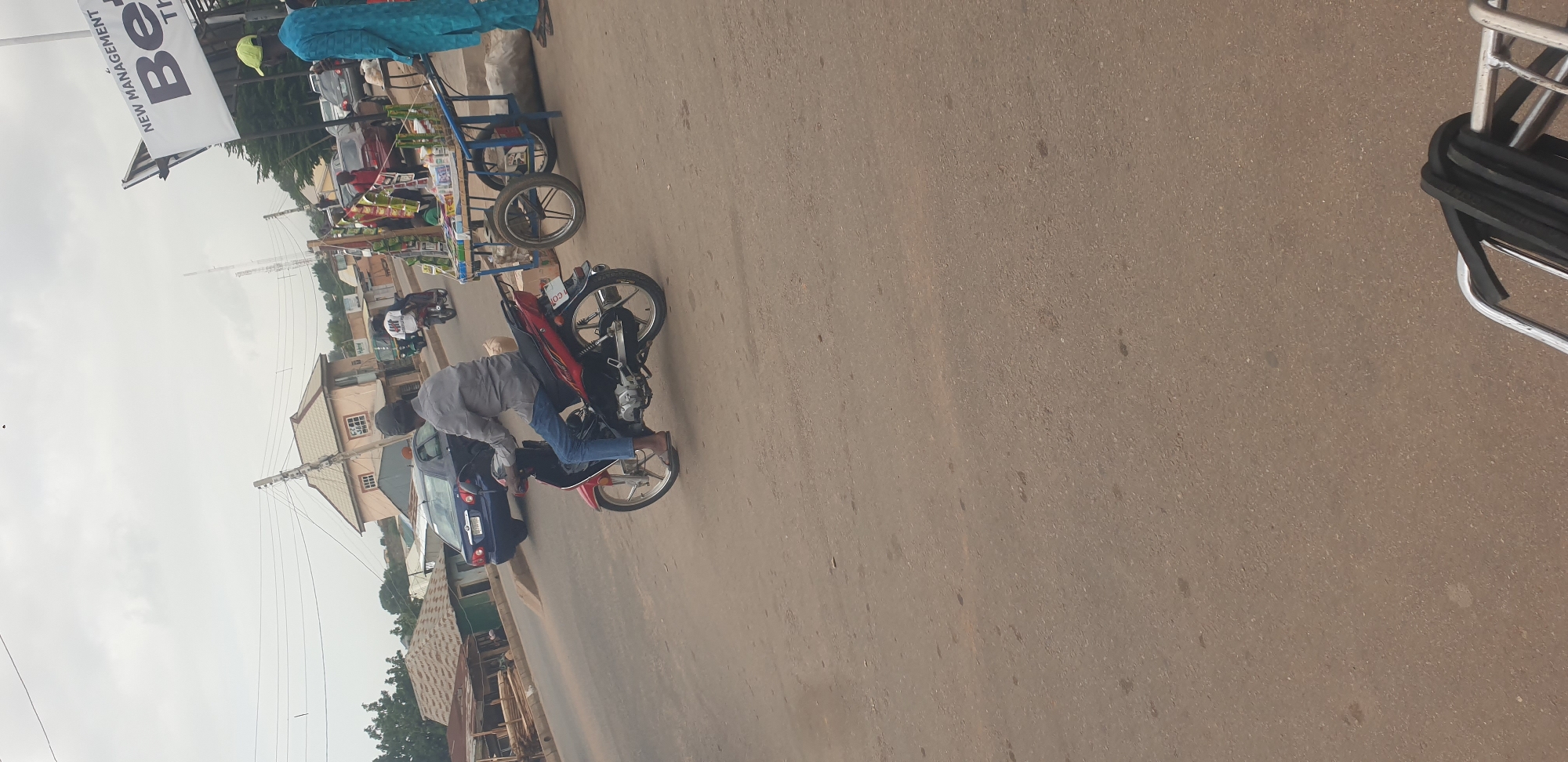 The image depicts a street scene in Barakin Lerawi, Nigeria. The main subjects are a man on a red motorcycle, leaning over it as if about to ride or having just stopped, and a street vendor's cart laden with various items, including bottled drinks and packaged goods. The setting appears to be a roadside market or a busy street with buildings and other vehicles, including a dark blue car, visible in the background.
The activity taking place is primarily the movement of people and goods in an urban environment. The man on the motorcycle seems to be a local commuter or a delivery person. The vendor's cart suggests commerce and daily life in the area.
Notable details include the overcast sky, indicating potentially cloudy weather. The time of day is difficult to determine precisely but appears to be daytime. The buildings in the background have a typical architectural style for the region. The man on the motorcycle is wearing a gray long-sleeved shirt, blue jeans, and a helmet. The vendor, only partially visible, is wearing a blue patterned garment.
Visible text includes "Be" and "NEW MANAGEMENT" on a white banner at the top left of the image, likely advertising a business. A license plate on the motorcycle is partially visible with "C01" printed on it. The overall mood is one of everyday hustle and bustle.
