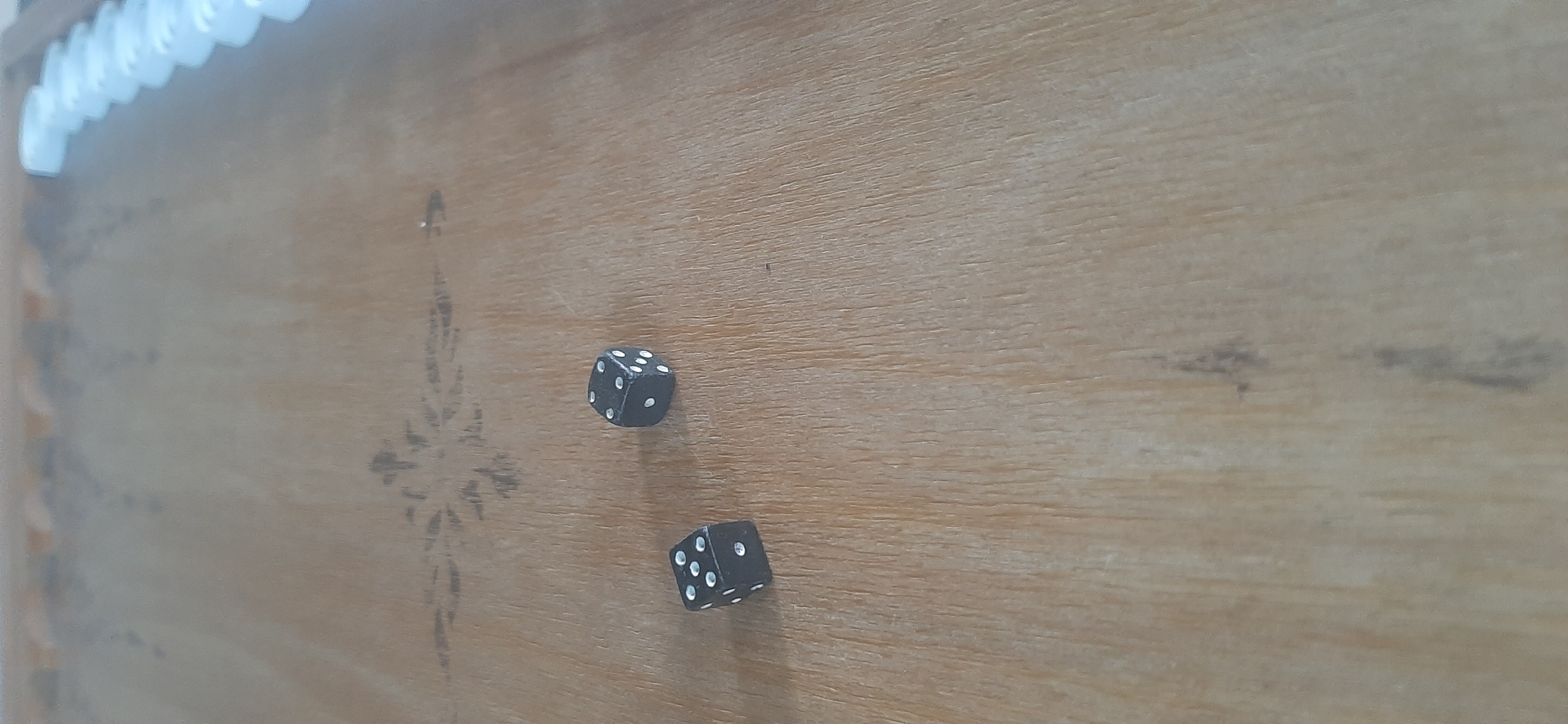 Two black dice with white pips are lying on a wooden surface. The die on the left shows a 5, and the die on the right shows a 3. The wooden surface appears to be a tabletop or a board, possibly for a game. There are some faint decorative patterns on the surface, and a blurred row of white objects in the background that could be game pieces. There are no people or animals present in the image. The scene is indoors, with natural light illuminating the surface. There is no information in the image to determine its location in Yessentukskaya, Russia.