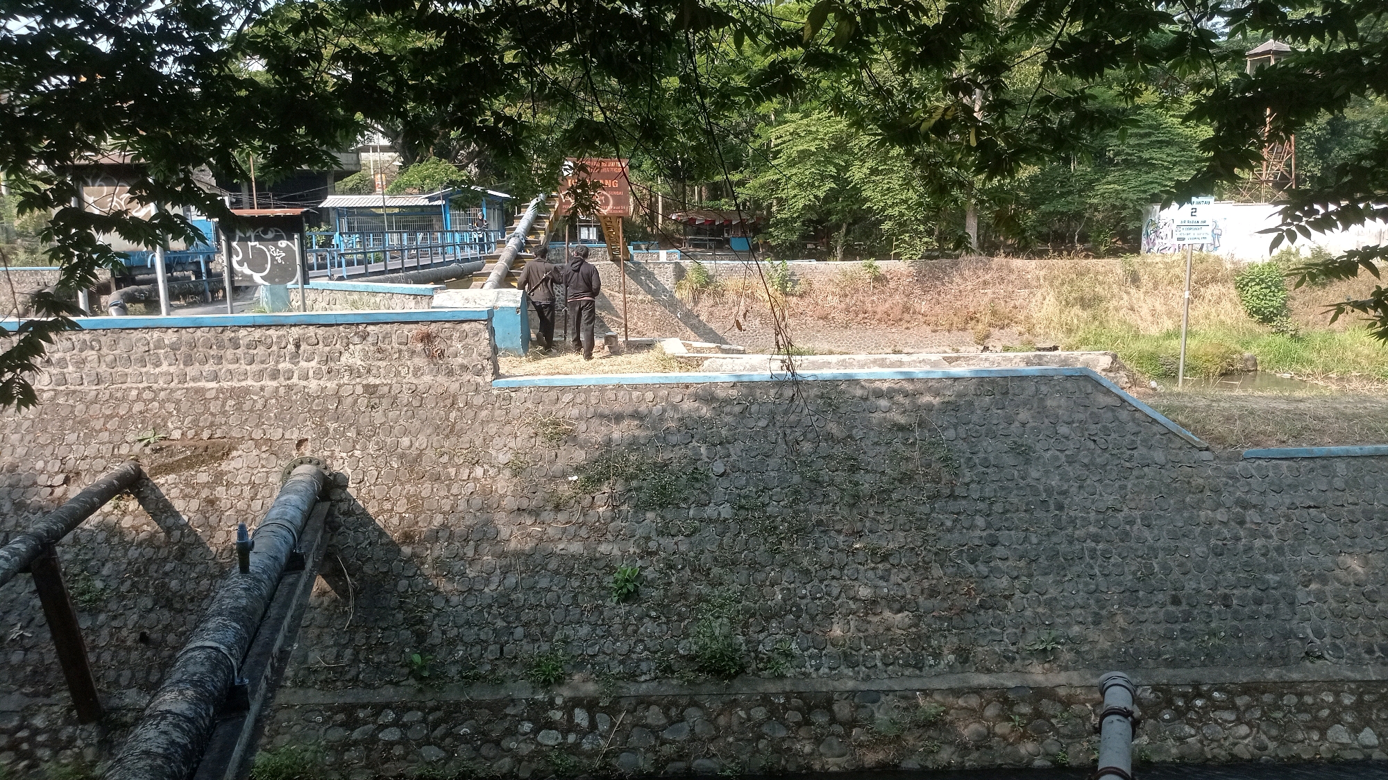 This image depicts a section of the Mulyoagung water treatment facility in Mulyoagung, Indonesia. The scene is dominated by a sturdy, stone-faced retaining wall. Two individuals, dressed in dark clothing, are seen walking away from the camera on a walkway atop the wall. Adjacent to them is a blue-railed structure, possibly a viewing platform or part of the facility's infrastructure, with some blue pipes visible. In the background, trees and vegetation form a natural backdrop, with glimpses of buildings and what appears to be a water tower further in the distance. Large diameter pipes are prominently visible in the foreground, running along the base of the retaining wall, indicating the presence of significant water flow or management systems. The overall impression is one of an industrial, yet natural, environment.