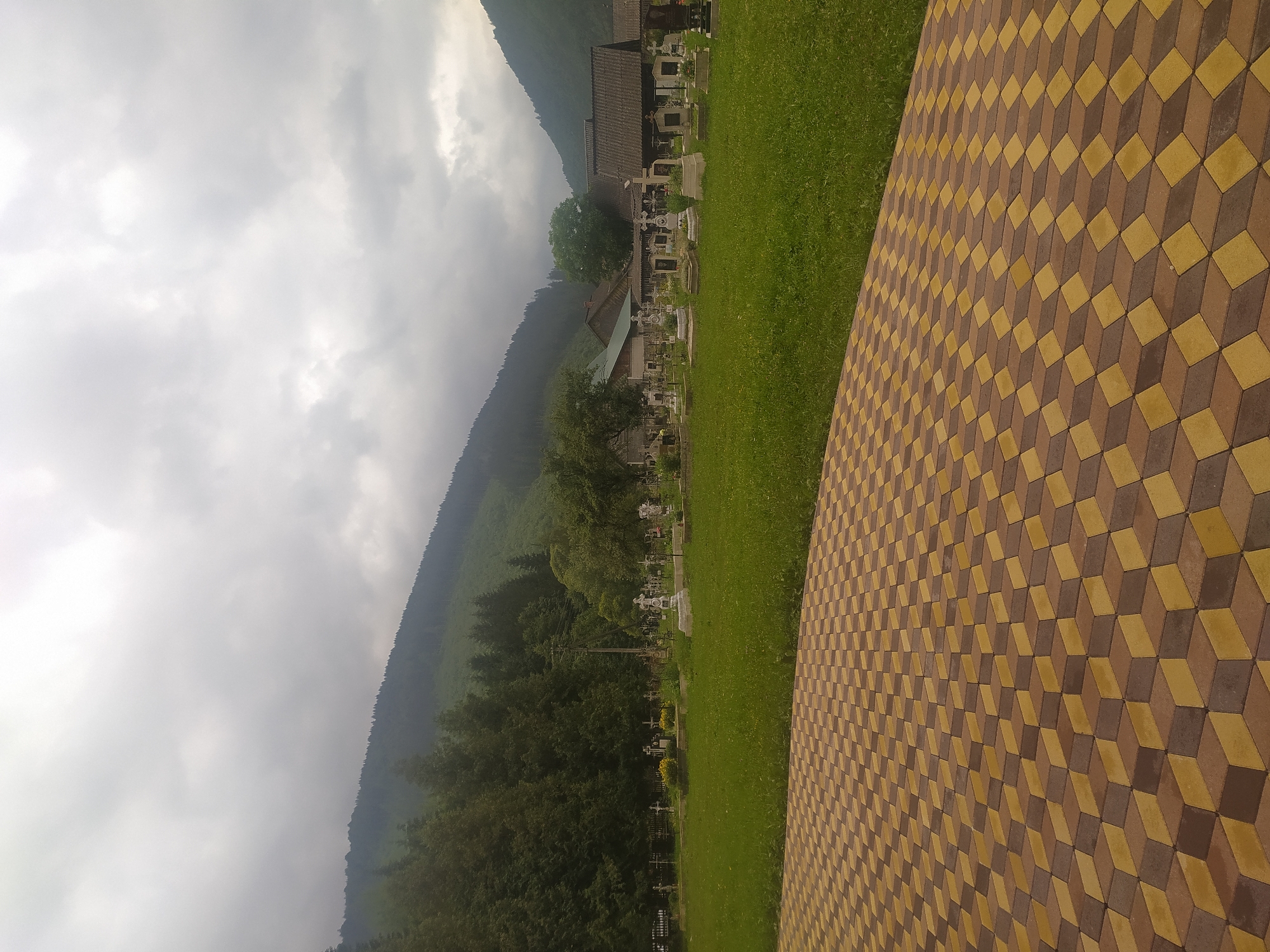 A cemetery is visible in the foothills of Luhy, Ukraine, with a cloudy sky overhead. The cemetery is filled with numerous gravestones and crosses of various sizes and designs, interspersed with green grass and trees. In the background, the landscape rises into forested hills. In the foreground, a patterned walkway made of brown and yellow interlocking bricks leads the eye into the scene. Several rustic buildings with wooden roofs are situated behind the cemetery.