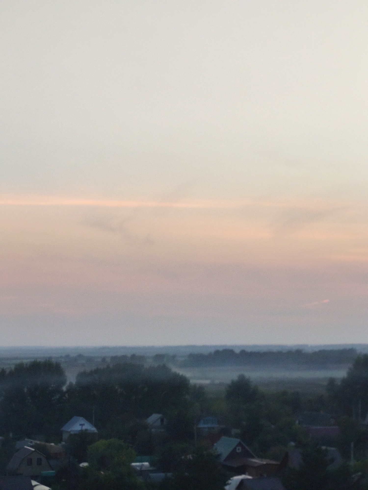 The image shows a landscape view with a hazy sky that transitions from a pale yellow at the top to soft pink and then a light gray towards the horizon. Below the sky, a dark band of trees and buildings stretches across the middle ground. In the foreground, there are several structures, including houses with pitched roofs, some partially obscured by foliage. The overall impression is of a rural or suburban area at dusk or dawn, with a low-lying mist or fog present in the distance. There are no people visible in the image. The scene depicts a quiet, possibly undeveloped area within the city of Petropavl, Kazakhstan, characterized by its natural elements and scattered residential buildings.