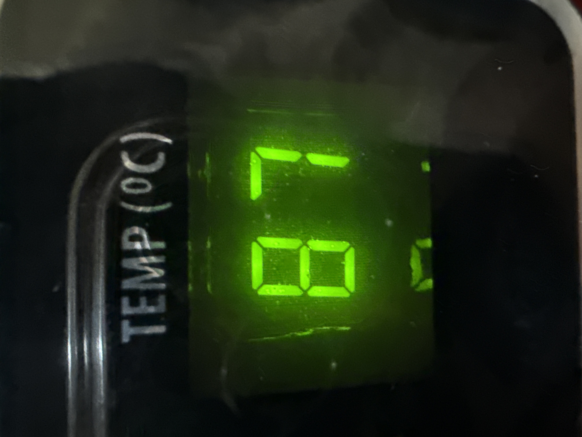 A close-up, high-angle shot shows a digital display with the text "TEMP (°C)" above a numerical reading. The numbers "8" and "7" are prominently displayed in a bright green, seven-segment digital format. The surrounding area is dark and appears to be part of an appliance or control panel. The image was taken in Lhokseumawe, Indonesia.
