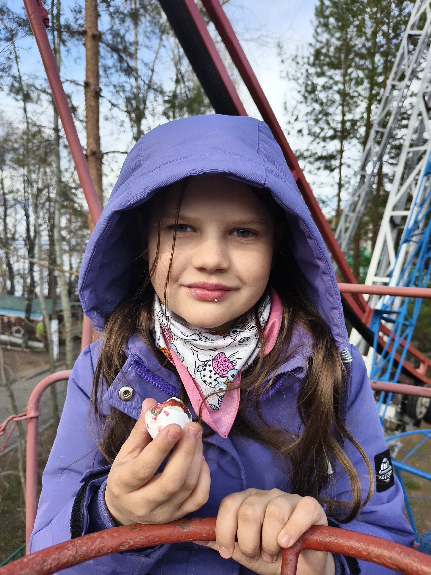 A young girl is featured in a close-up, chest-up portrait, looking directly at the camera with a slight smile. She wears a vibrant purple hooded jacket, with the hood pulled up, and a white scarf with colorful patterns and a pink trim around her neck. Her long, dark hair frames her face, and she has visible freckles across her nose and cheeks. A small silver labret piercing is visible on her lower lip.
In her left hand, she holds a small, white object with red markings, possibly a decorated egg or a piece of a patterned shell. Both her hands are resting on a weathered, red metal bar, suggesting she is seated on or interacting with a structure.
The background is out of focus but reveals an outdoor setting, likely an amusement park or fairground. Prominent red metal bars are close behind her, and further back, there are larger, intricate metal structures in shades of blue, white, and red, typical of amusement park rides such as rollercoasters or Ferris wheels. Bare trees and an overcast sky are visible between the structures. A small, dark emblem or patch is present on the upper sleeve of her right arm, but any text or design on it is indistinct. The lighting suggests it is daytime, possibly under an overcast sky.