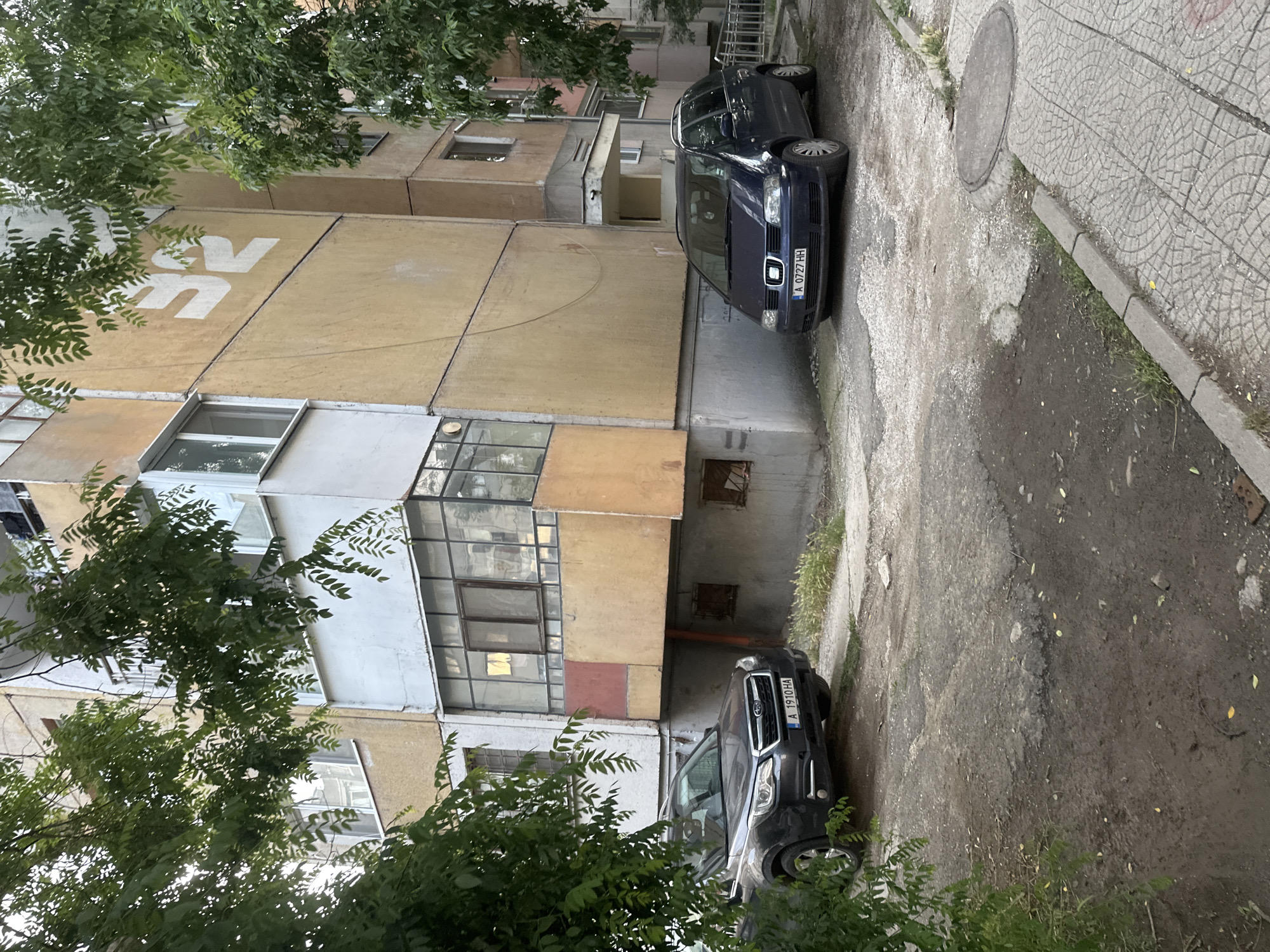 The image presents an outdoor residential scene in Burgas, Bulgaria, featuring a multi-story apartment building, two parked cars, and substantial greenery. The setting appears to be an urban or suburban street on an overcast day.
On the left, a large, worn apartment building with a yellowish-beige facade dominates the scene. Its exterior shows signs of age, with some peeling paint and visible panel lines. Various windows and enclosed balconies are present on different floors. A prominent white graphic, resembling stylized letters "SV" or "SN," is painted on the upper part of the building's facade, partially obscured by tree branches.
Two cars are visible. A dark blue sedan, possibly a Seat model, is parked closer to the building on the left side of a dirt patch. Its Bulgarian license plate reads "A 0727 HH." Further down and to the right, a dark SUV, possibly a Subaru, is partially visible, also parked on the dirt area. Its license plate is "A 1910 HA." Both license plates begin with 'A', indicating the Burgas region.
The ground consists of a mixed surface: a dirt and gravel patch where the cars are parked, and a paved sidewalk with a decorative circular pattern on the right. A metal manhole cover is set into the sidewalk. Lush green trees with dense foliage are abundant, framing the scene from the top and bottom left, casting dappled shadows, and partially obscuring parts of the buildings and vehicles. No people or specific activities are observable, suggesting a quiet, static moment in a residential area. The overall impression is one of everyday urban life in Bulgaria.