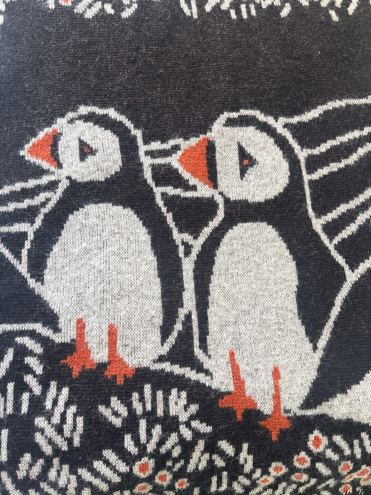 This is a close-up, flat-lay photograph of a knitted textile featuring a repeating pattern of two puffins with orange beaks and feet, set against a dark background. White lines create decorative elements around the puffins, resembling foliage or waves. The puffins are depicted in profile, with their bodies in white and black, and their faces detailed with white patches and orange triangles for beaks. The textile appears to be a blanket or fabric, with visible texture from the knitting. There is no indication of Reykjavik, Iceland, in the image itself.