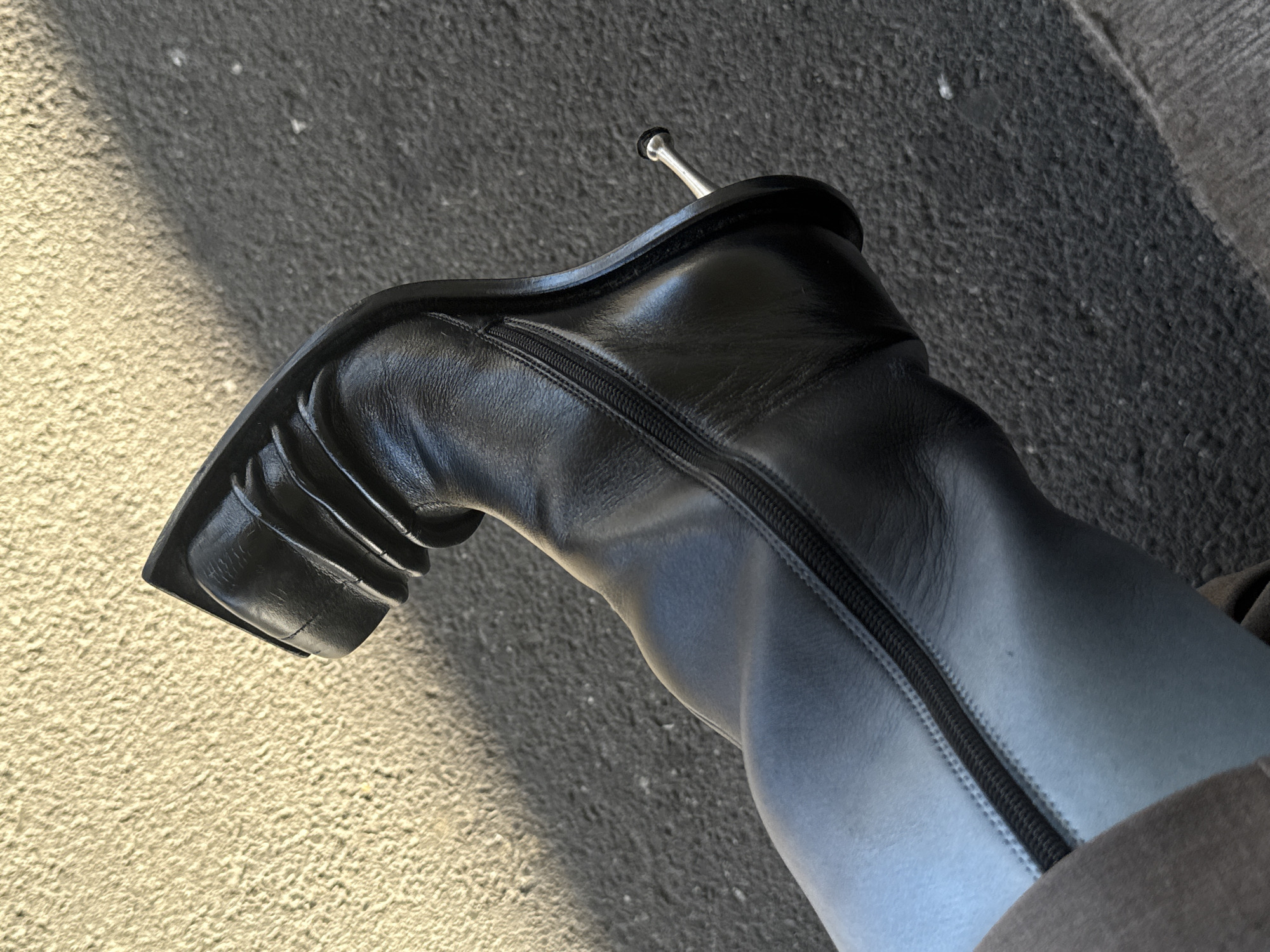 This close-up, angled shot features a person's leg encased in a sleek, black leather boot. The boot has a distinctive square toe, a tall, block-like heel, and a metallic, silver-colored pin or tip protruding from the very bottom of the heel. A black zipper runs vertically along the side of the boot. The leg appears to be covered by dark, form-fitting trousers or tights, blending seamlessly with the boot's upper.
The setting is outdoors, with the boot resting on a textured ground surface. The foreground consists of a light, yellowish-beige, rough-textured pavement or concrete, while the background transitions to a darker, greyish-black asphalt or similar ground material. A distinct shadow line separates these two areas, suggesting strong natural light from an unseen source, indicating daytime. The scene suggests a static pose or a momentary stop during a walk in an urban environment. No specific location cues are visible in the image itself, though the context provided specifies London, United Kingdom. There are no other people visible, no discernible emotions conveyed, and no visible text. The image serves as a detail shot, highlighting the footwear.