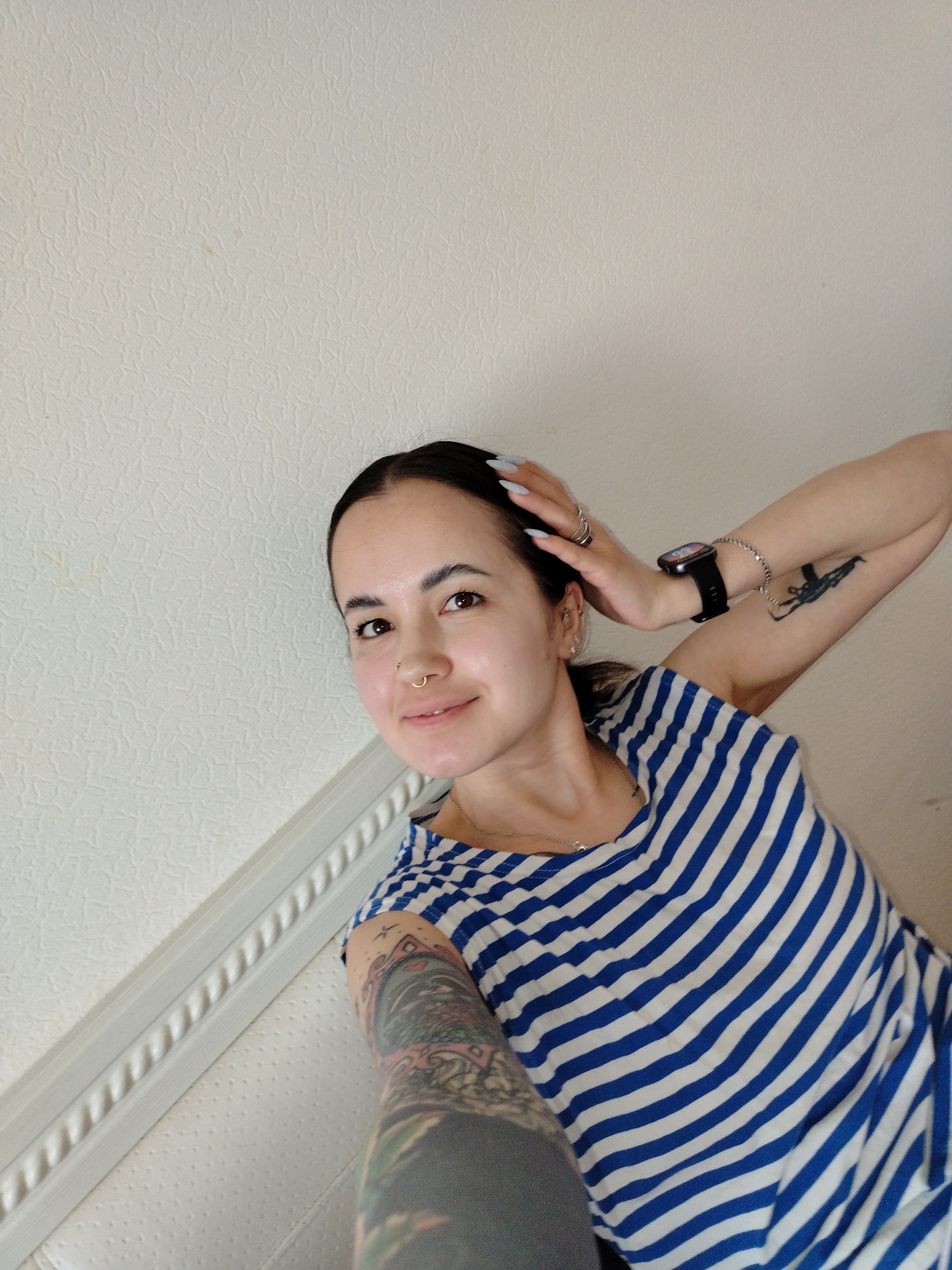 A young woman is captured in a selfie-style shot, leaning against a textured, light-colored wall indoors, likely in Noviy, Russia. She has dark hair pulled back, brown eyes, and a subtle smile, looking directly at the camera. A small gold hoop nose piercing adorns her right nostril.
She is wearing a blue and white striped top, reminiscent of a marinière. Her left arm is raised, with her hand touching the back of her head, showcasing light blue, long fingernails, multiple rings, a black smartwatch, and a silver bracelet. A distinct black silhouette tattoo of a rifle, resembling an AK-47, is visible on her upper left arm. Her right arm, extending towards the camera, is adorned with a colorful, intricate full-sleeve tattoo. A silver necklace with a small pendant is also visible around her neck, and she has small earrings in both ears.
The background consists of a uniformly light beige or off-white wall with a subtle, irregular textured pattern. In the lower left, a white decorative molding with a rope-like design is present, possibly part of a bed frame or wainscoting. The lighting appears soft and natural, suggesting daytime. No discernible text is visible in the image. The overall impression is a casual, personal photograph.