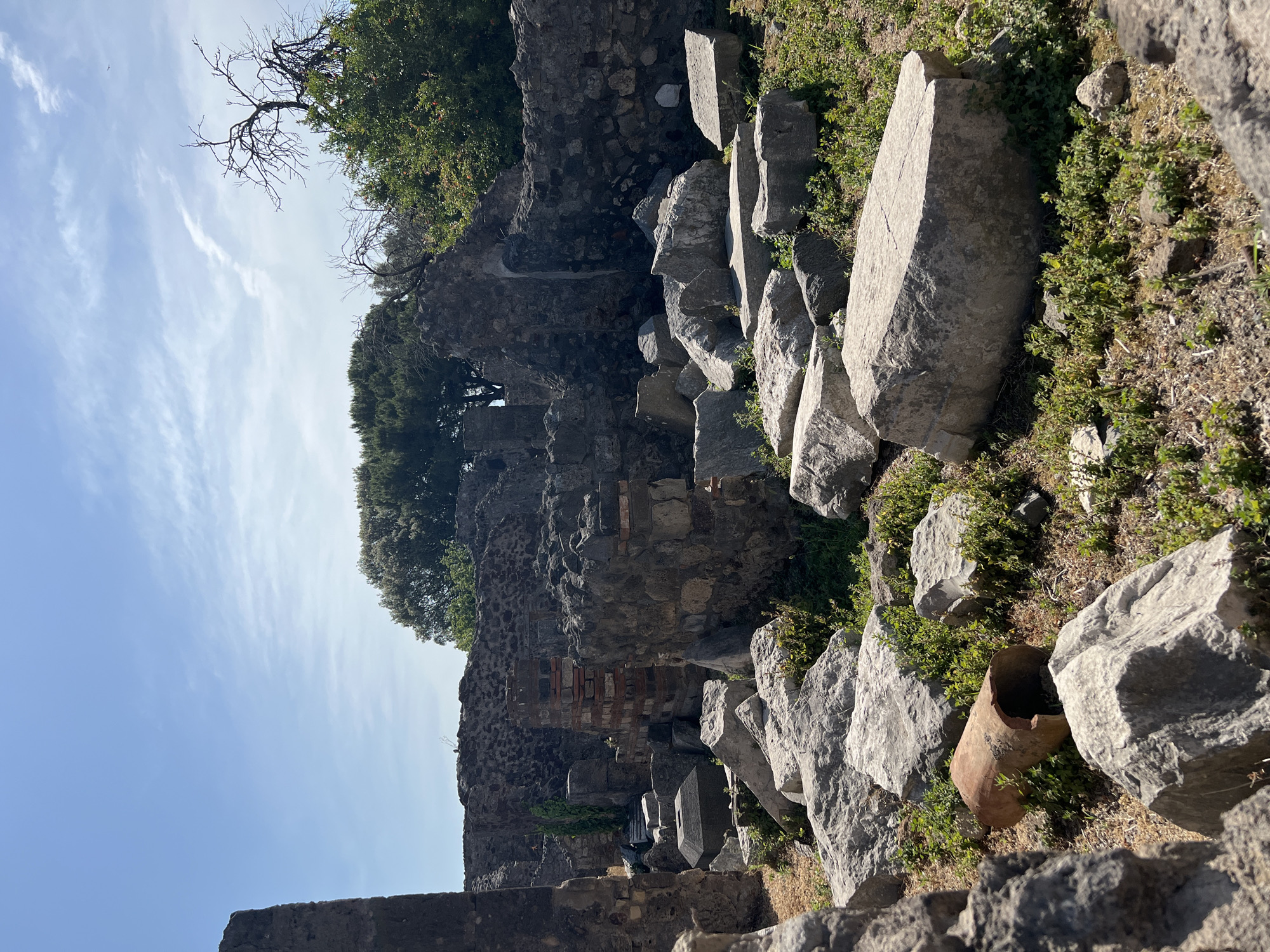 The image shows ruins in Pompeii, Italy, under a bright blue sky with wispy clouds. The foreground is filled with large, weathered grey stones, some appearing to be architectural fragments, scattered amongst green vegetation and dry earth. A broken terracotta pipe lies horizontally amongst the stones in the mid-ground. Behind the stones, remnants of stone walls and brick structures rise, some partially crumbled. Overgrown trees and vegetation are visible in the background, partially obscuring the sky. The scene depicts an archaeological site with evidence of ancient construction and natural overgrowth.