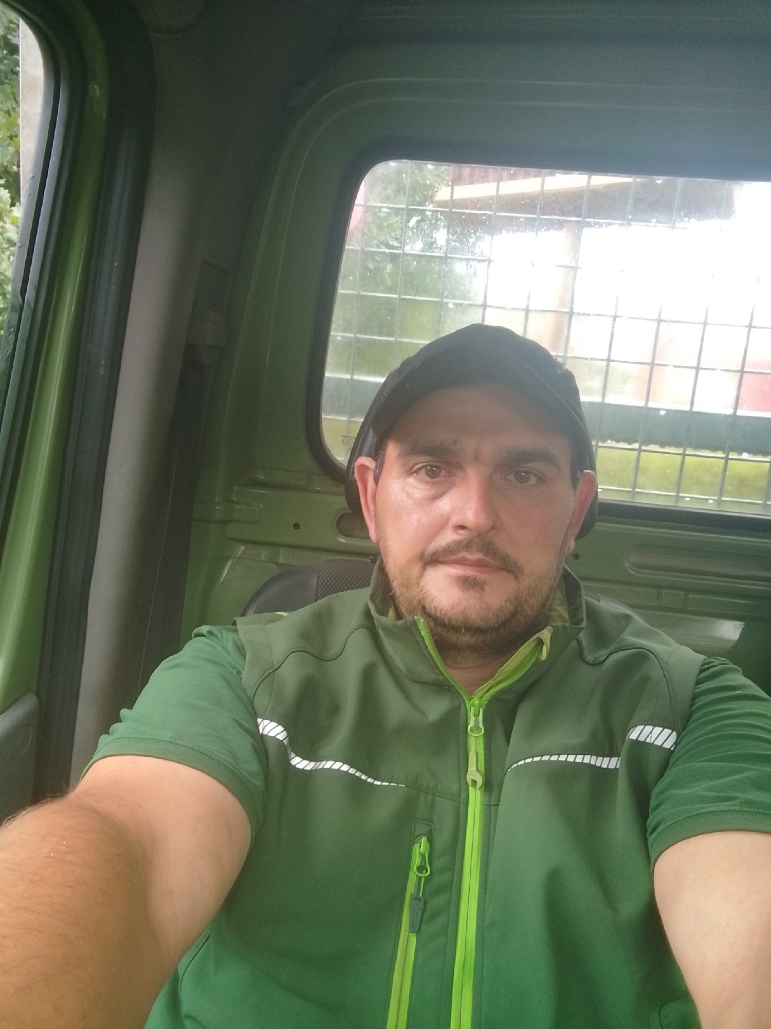 A man wearing a green shirt and a green vest with a black baseball cap is sitting inside a green vehicle, likely a truck cab. He is looking at the camera and has a mustache and beard. The vehicle's interior is visible, with green paneling and a window behind him. The window has a metal grid and shows an outdoor scene with trees and some structures, possibly indicating a rural or semi-rural location. The lighting suggests it is daytime. The image does not provide any specific clues to pinpoint a location within the city of Hamburg, Germany.