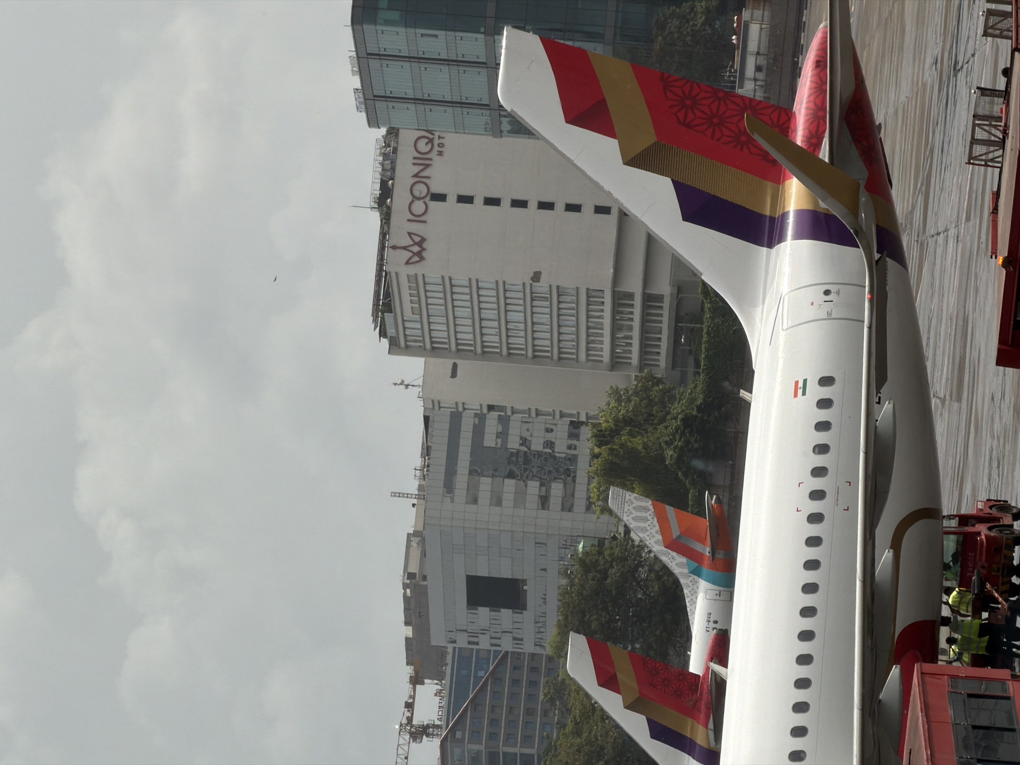 The image depicts an aerial view of aircraft parked on a tarmac, with several modern buildings and a construction crane visible in the background. The planes have distinctive tail fin designs in vibrant colors, including red, gold, purple, and teal, with intricate patterns. The largest aircraft in the foreground is primarily white, with windows lining its fuselage. A red baggage tug with workers in high-visibility vests is positioned beneath the front of this aircraft.
In the middle ground, two other aircraft tails are visible, one with a teal and orange pattern and the other red and gold. Behind the aircraft, a row of trees separates the tarmac from a cluster of buildings. The most prominent building in the background is a multi-story structure with a white facade, bearing the large red logo "ICONIQ" and "HOT" on its upper section. To the right of this is a modern glass-fronted building. The sky is overcast with heavy clouds. The scene is consistent with an airport environment, likely within the Mumbai Metropolitan Region, given the architectural style of the buildings and the presence of the "ICONIQ" hotel, which is located in Mumbai.