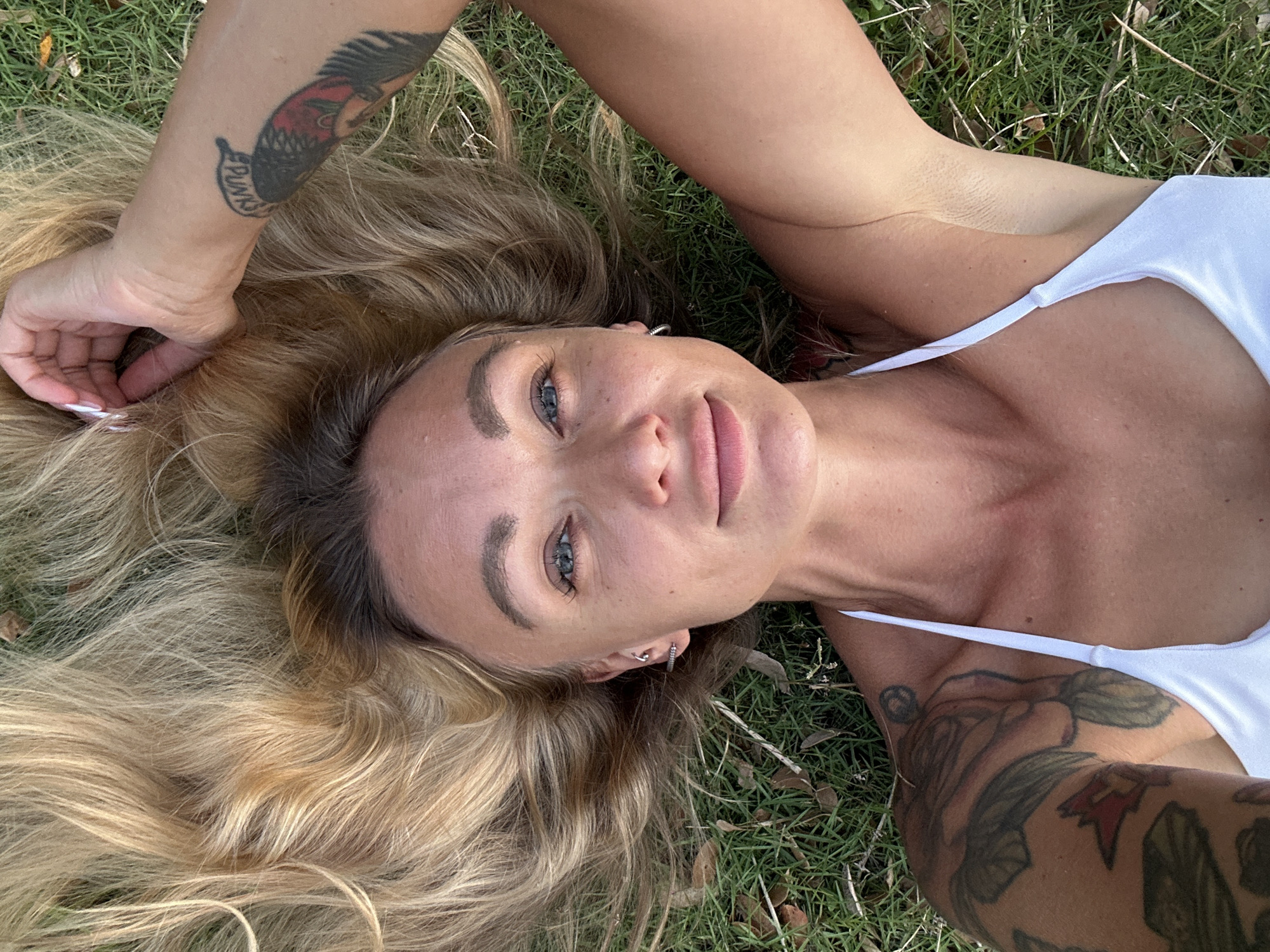 A woman with blonde hair lies on grass in Tien Thanh Ward, Vietnam. She is wearing a white top and has tattoos on her arms. Her left arm is raised and resting on her head, while her right arm is down by her side. The woman's face is partially visible, and she is looking up at the camera. The grass is green and appears to be well-maintained. There are a few fallen leaves scattered on the grass. The lighting suggests it is daytime, with the sun casting subtle shadows.