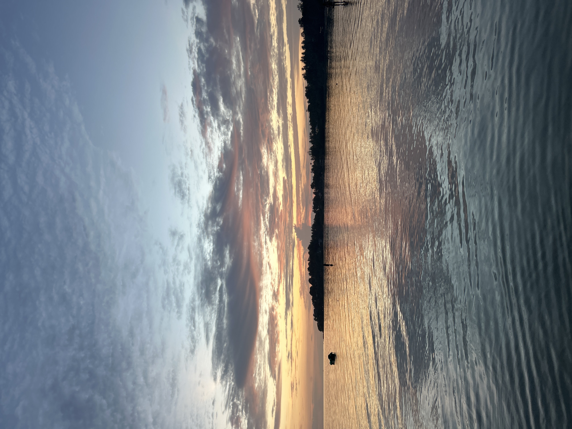 An expansive body of water reflects the warm hues of a sunset, casting orange and pink tones across its surface. The horizon is marked by a dense line of trees, silhouetted against the sky. The sky itself is a canvas of blues and grays, interspersed with streaks of pink and orange from the setting sun. A small boat rests on the water in the distance, and two figures are visible standing in the shallow water near the shoreline. The scene is serene, capturing the tranquil beauty of a coastal evening in Ko Pha-ngan District, Thailand.