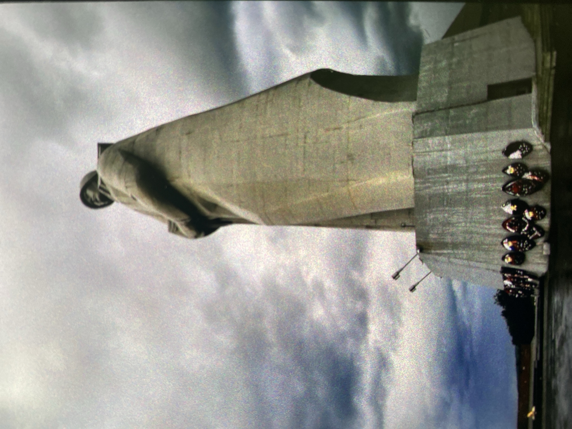 The image depicts the Motherland Monument in Saint Petersburg, Russia, from a low angle. The overcast sky dominates the background. The statue is rendered in a gray concrete, with a visible base. There are numerous people at the base of the statue, and streetlights can be seen.