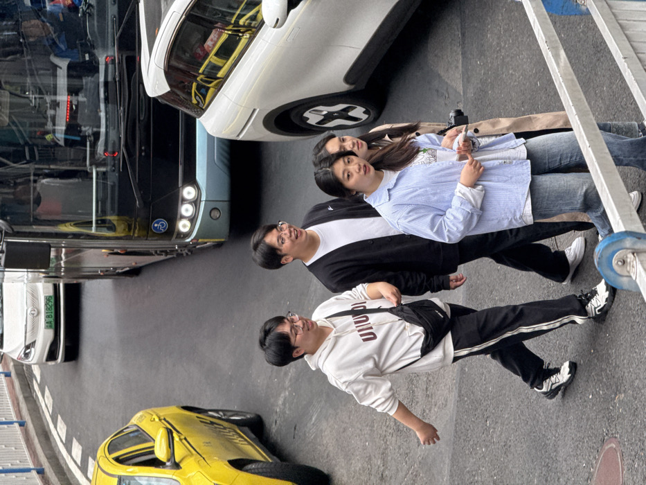 Four individuals are seen on a city street next to various vehicles, including a yellow sports car and buses. The group consists of two young women and two young men. The men are wearing glasses and casual clothing. One is in a white hoodie and black pants while the other is in a black jacket over a white top and black pants. The women are wearing jeans and light-colored tops. A ladder is visible in the foreground.