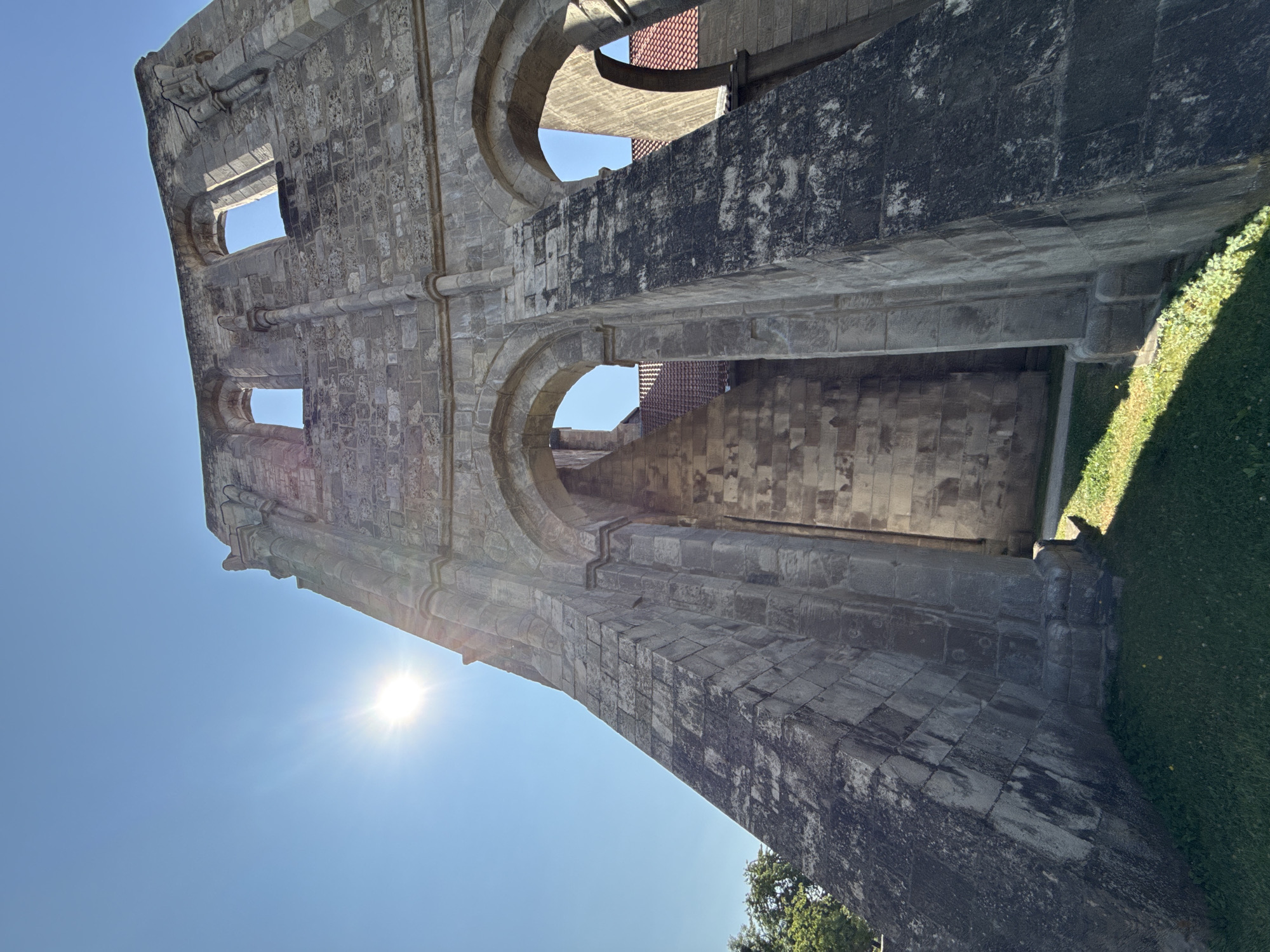 The image shows the ruins of Walkenried Abbey, located in Walkenried, Germany. The ruins are made of large, weathered stone blocks and feature arched openings. The sun is shining brightly in a clear blue sky, casting shadows on the ground. The foreground consists of green grass with some patches of yellow flowers. Trees are visible on the left side of the image. The architecture suggests a historical religious building, likely a church or monastery.