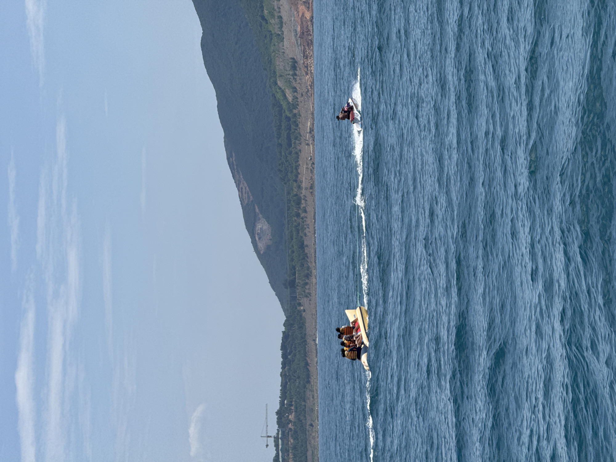 The image depicts a scene at sea near Nha Trang, Vietnam, under a bright, partly cloudy sky. In the foreground, the deep blue ocean is choppy with small waves. Two recreational watercraft are in motion. On the left, a yellow banana boat is being towed, carrying four individuals wearing life vests. On the right, a person is riding a jet ski, creating a white wake behind them. In the background, a distant shoreline features a lush green forested mountain range. A construction crane is visible on the left side of the distant landscape, indicating ongoing development. The overall atmosphere is one of a sunny day at a coastal recreational area.