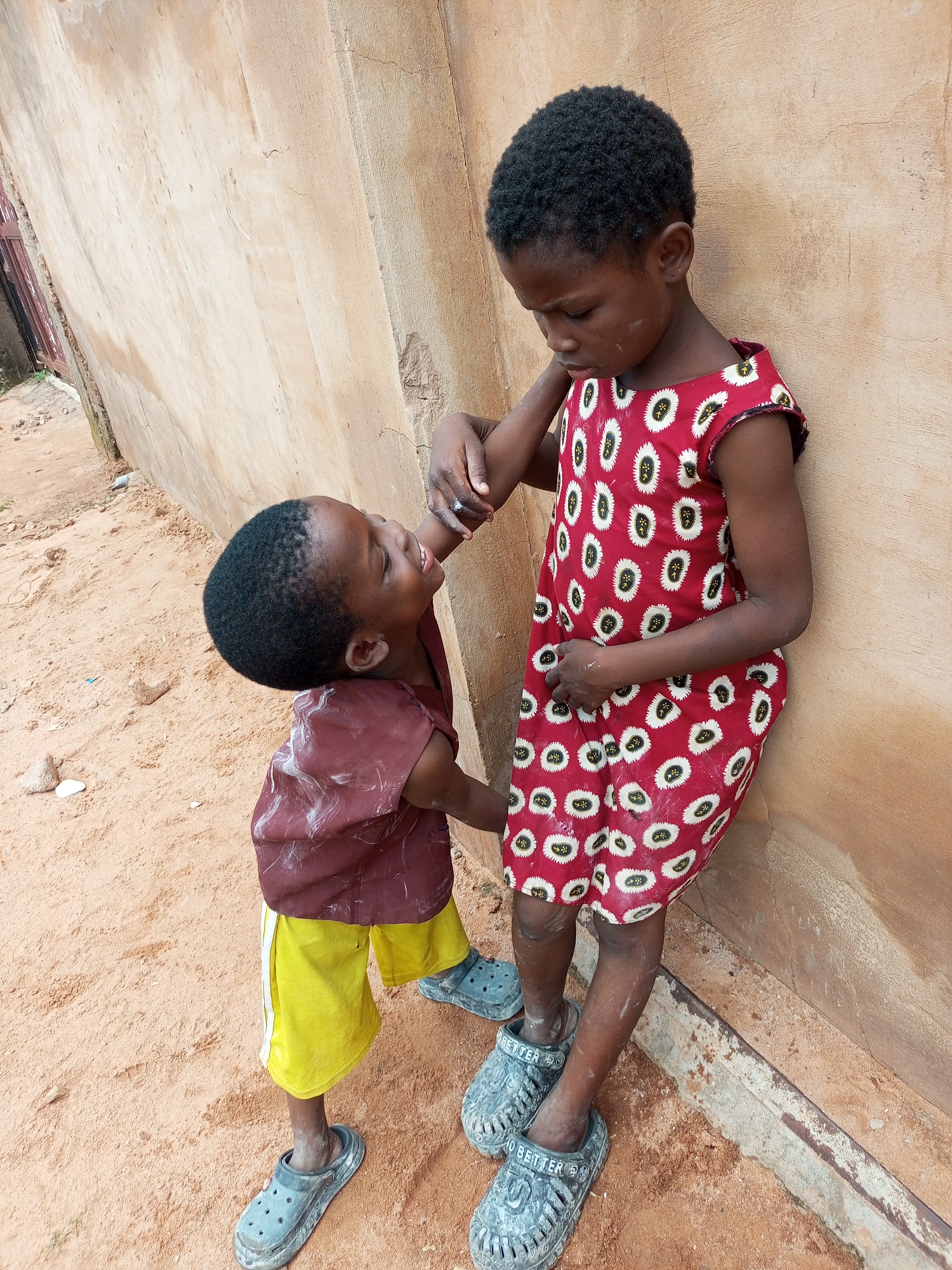 Two young children, a boy and a girl, stand outside next to a light brown stucco wall in Tempure, Nigeria. The girl, taller of the two, wears a red dress with a white and black pattern and stands leaning against the wall. The boy, smaller and facing the girl, wears a maroon shirt and yellow shorts. He is looking up at the girl and holding her hand. Both children are wearing grey slip-on shoes. The ground is covered in orange sand, with some small stones visible. The scene appears to be a residential area within the city.