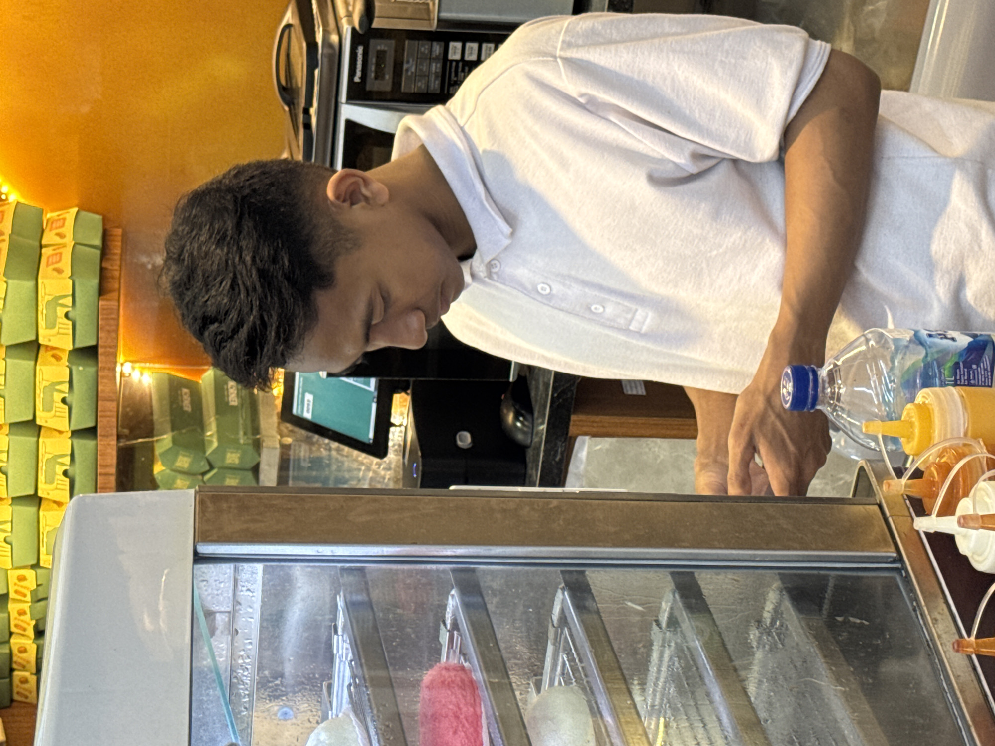 A young Indonesian male, wearing a light-colored polo shirt, is standing in front of a glass display case filled with food items. He is looking down and appears to be engaged in an activity, possibly preparing or handling food. Behind him, shelves are stacked with green and yellow boxes, suggesting a retail or food service environment. A microwave oven and a computer monitor are visible in the background. In the foreground, several condiment bottles and a bottle of water are placed on a counter. The scene is lit with warm lighting, and the overall impression is that of a bustling food establishment in Kuta Selatan, Indonesia.