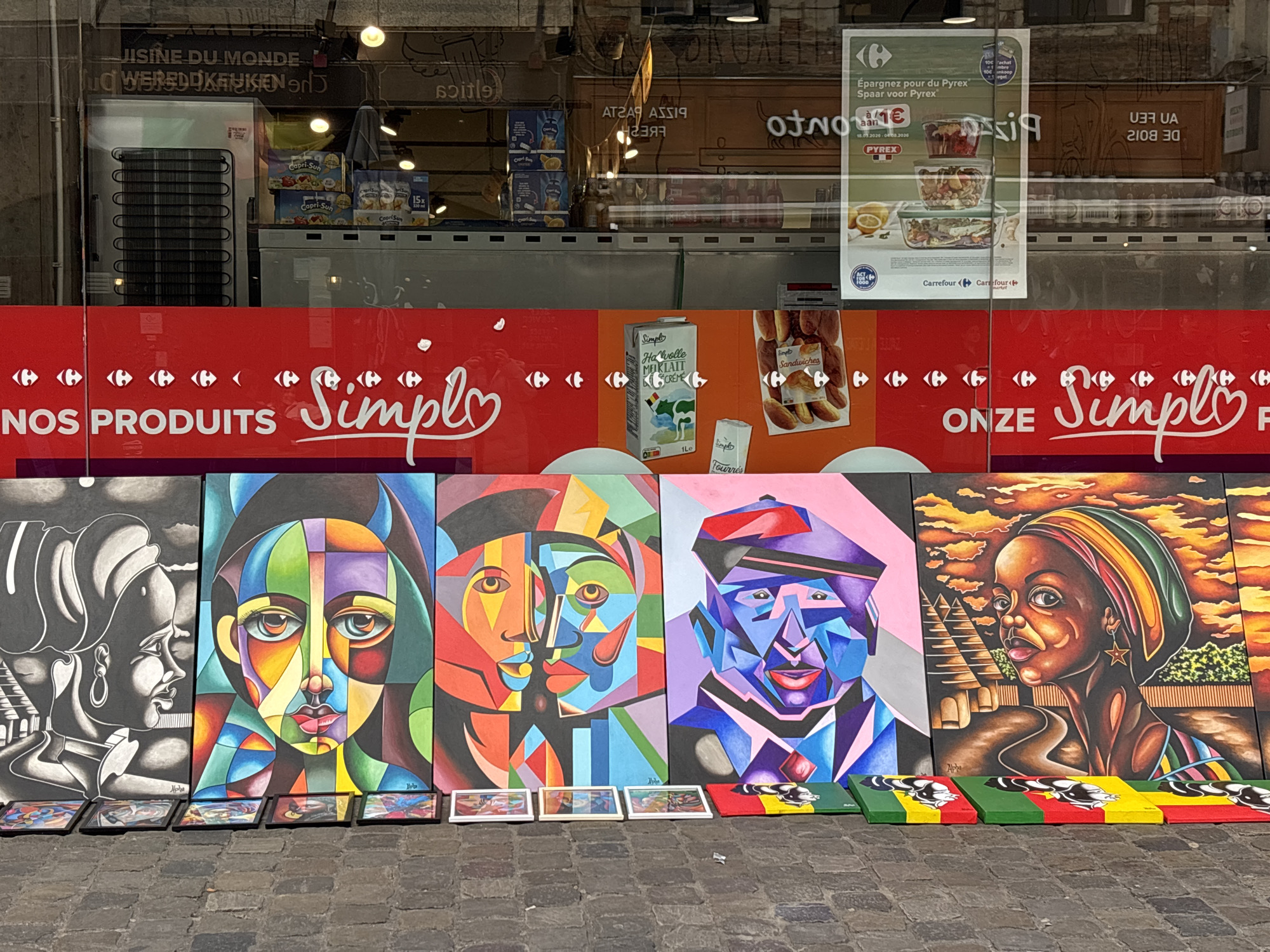 The image captures an outdoor display of paintings propped against the exterior wall of a Carrefour supermarket in Brussels, Belgium. The paintings depict abstract portraits with vibrant colors and geometric shapes. Below each painting are smaller reproductions or decorative pieces, some painted in the colors of the Beninese flag. The Carrefour's storefront features the "NOS PRODUITS Simply" branding. Various products are visible inside the store through the reflective windows, including food items like Capri-Sun juice boxes, Pyrex containers, milk, and sandwiches. Signs and posters, including an advertisement for a Pyrex promotion and the store's name in both French and Flemish ("UISINE DU MONDE" and "WEREDOKKEN"), are affixed to the glass. The ground is paved with cobblestones.