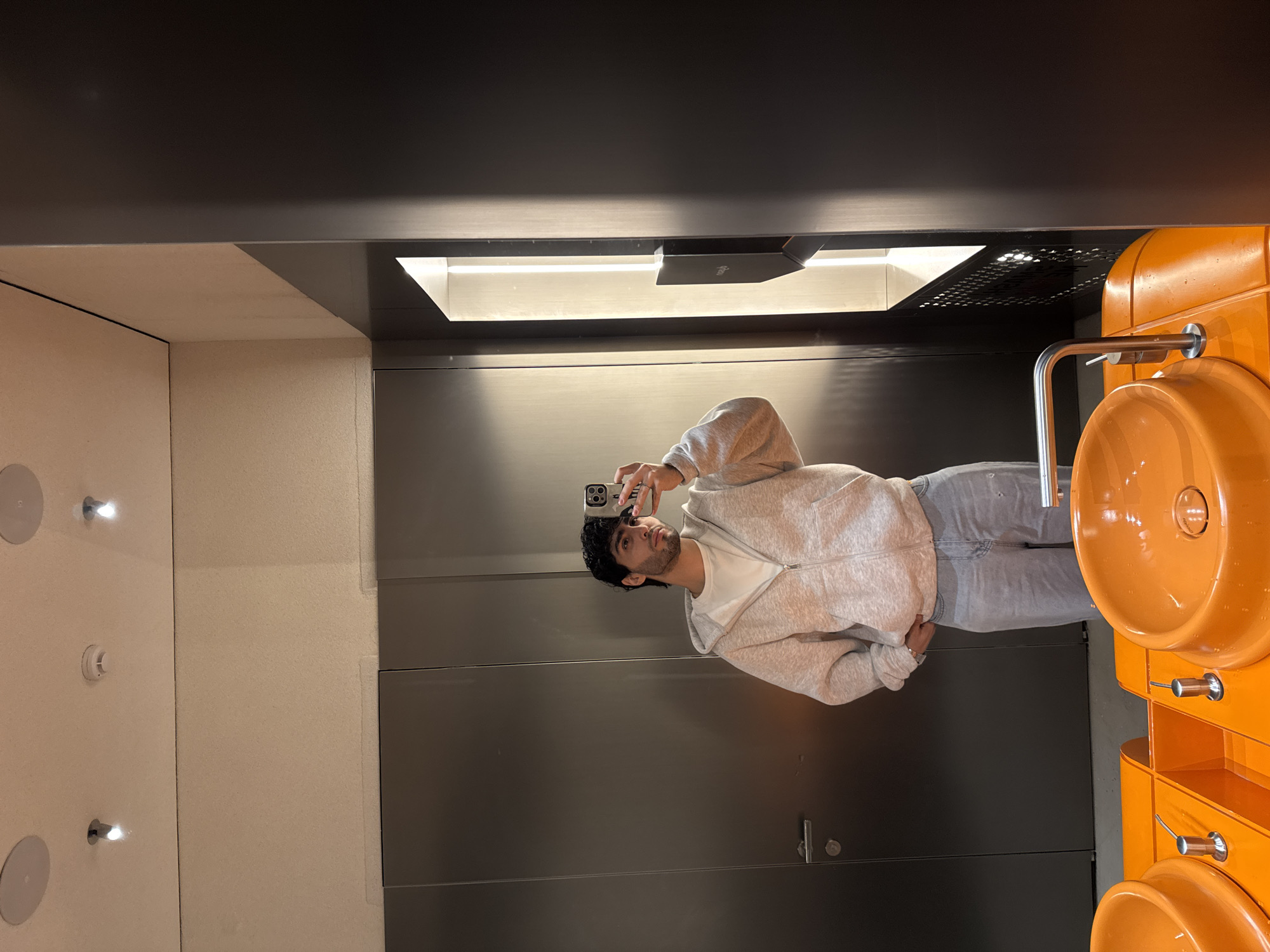 A man is taking a selfie in a bathroom. He is facing a dark gray wall with a door. To his right is an orange sink unit, and above him is a recessed light fixture. The opposite wall is beige with round ceiling lights. There is no specific information in the image to identify the location as Yerevan, Armenia.
