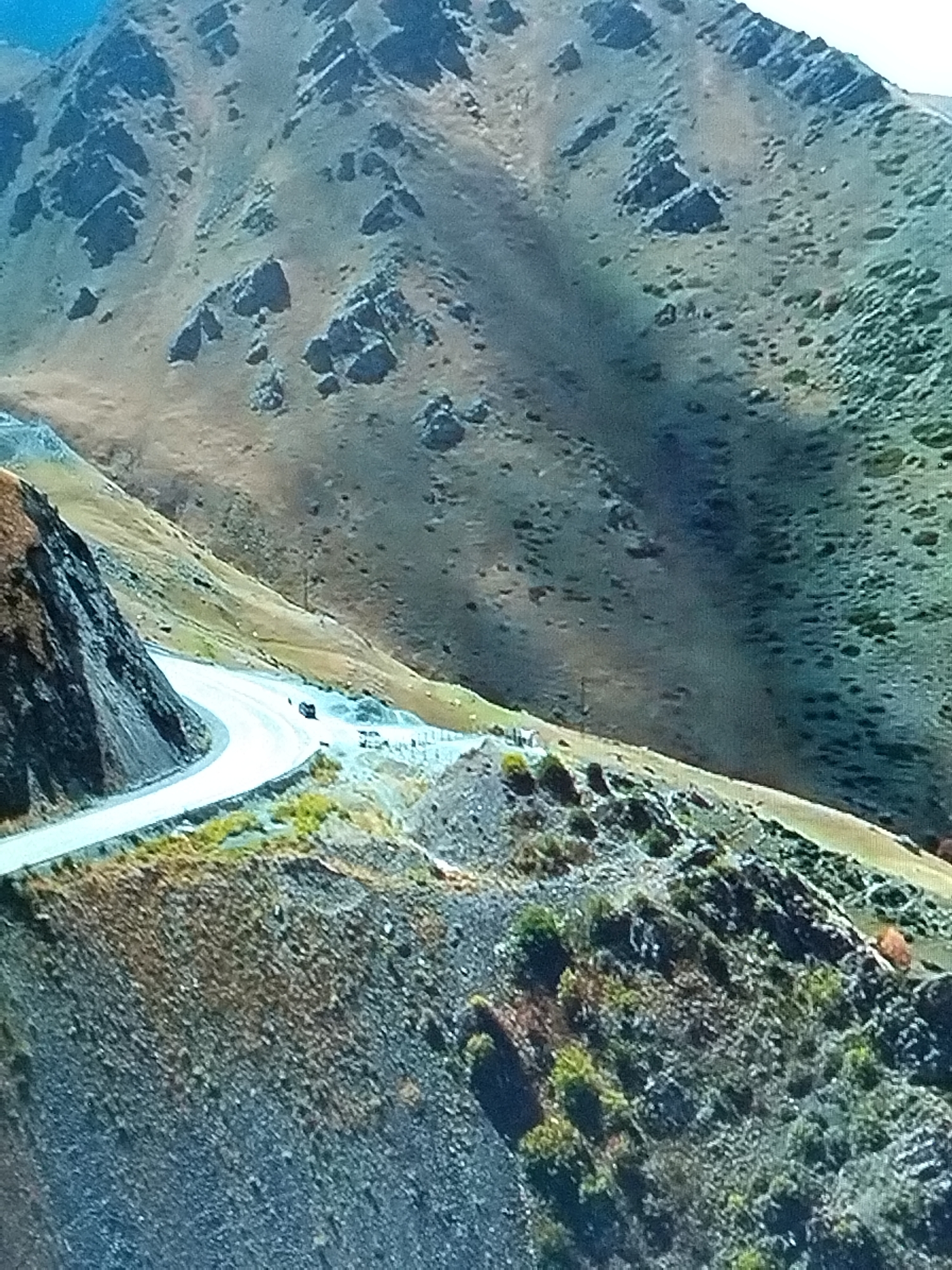 The image depicts a winding mountain road with a single dark-colored car driving on it. The road is paved and curves through a rugged, mountainous terrain. The landscape is characterized by steep slopes covered in sparse vegetation and rocky outcrops. The sky is partially visible at the top of the image and appears to be a bright blue. The overall scene suggests a remote, scenic location, likely in a mountainous region. There is no visible indication of any city or specific location.