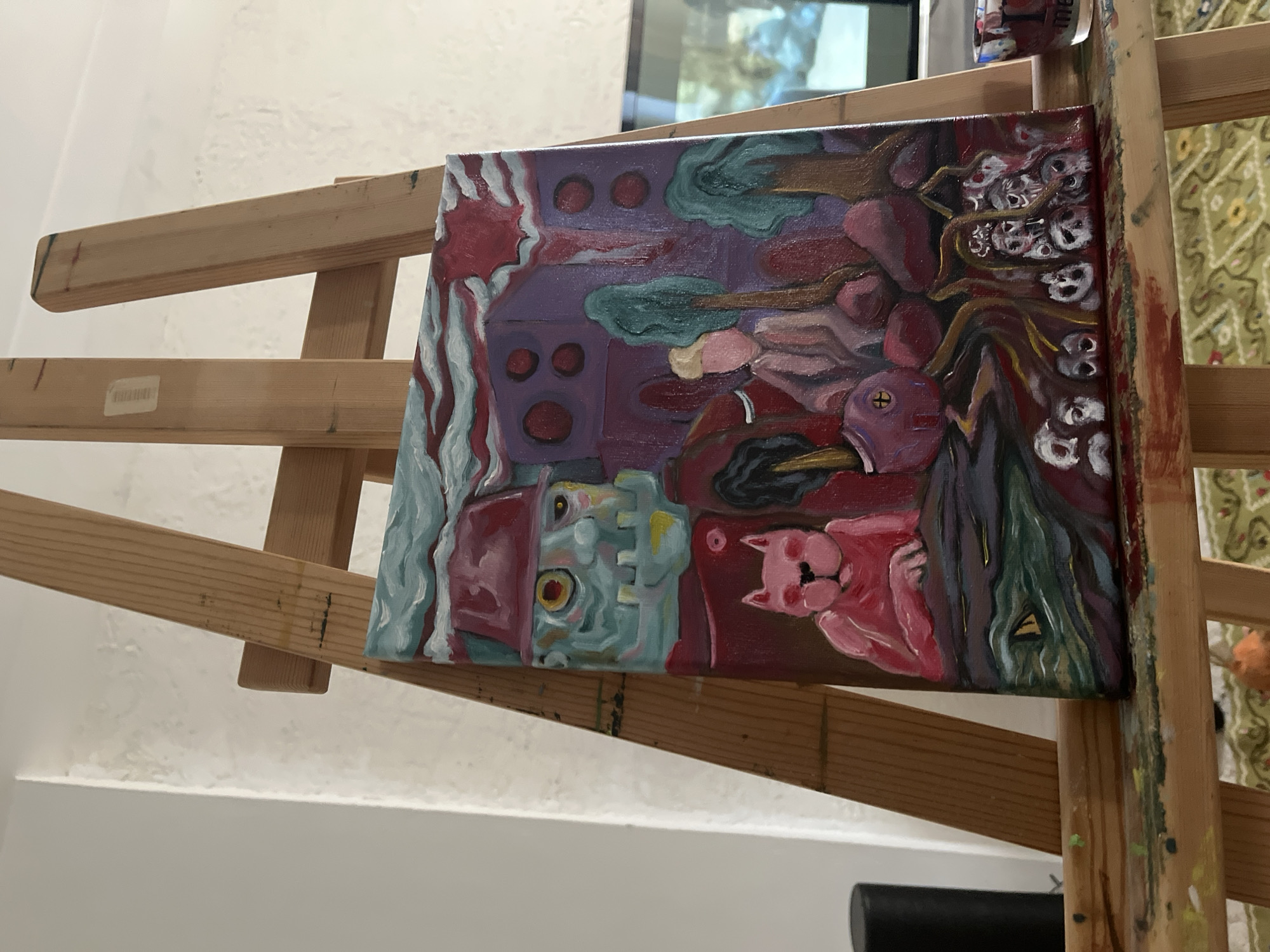 The image shows a painting on an easel. The painting is an abstract scene with a light blue figure in a red hat, a pink cat, and other shapes in various shades of red, purple, and green. The background of the painting includes a purple structure with red circles, green shapes that might be trees, and a group of small white skulls. The easel is made of light-colored wood and has paint splatters on it. The background is a white wall and a window with a view of trees and sky.