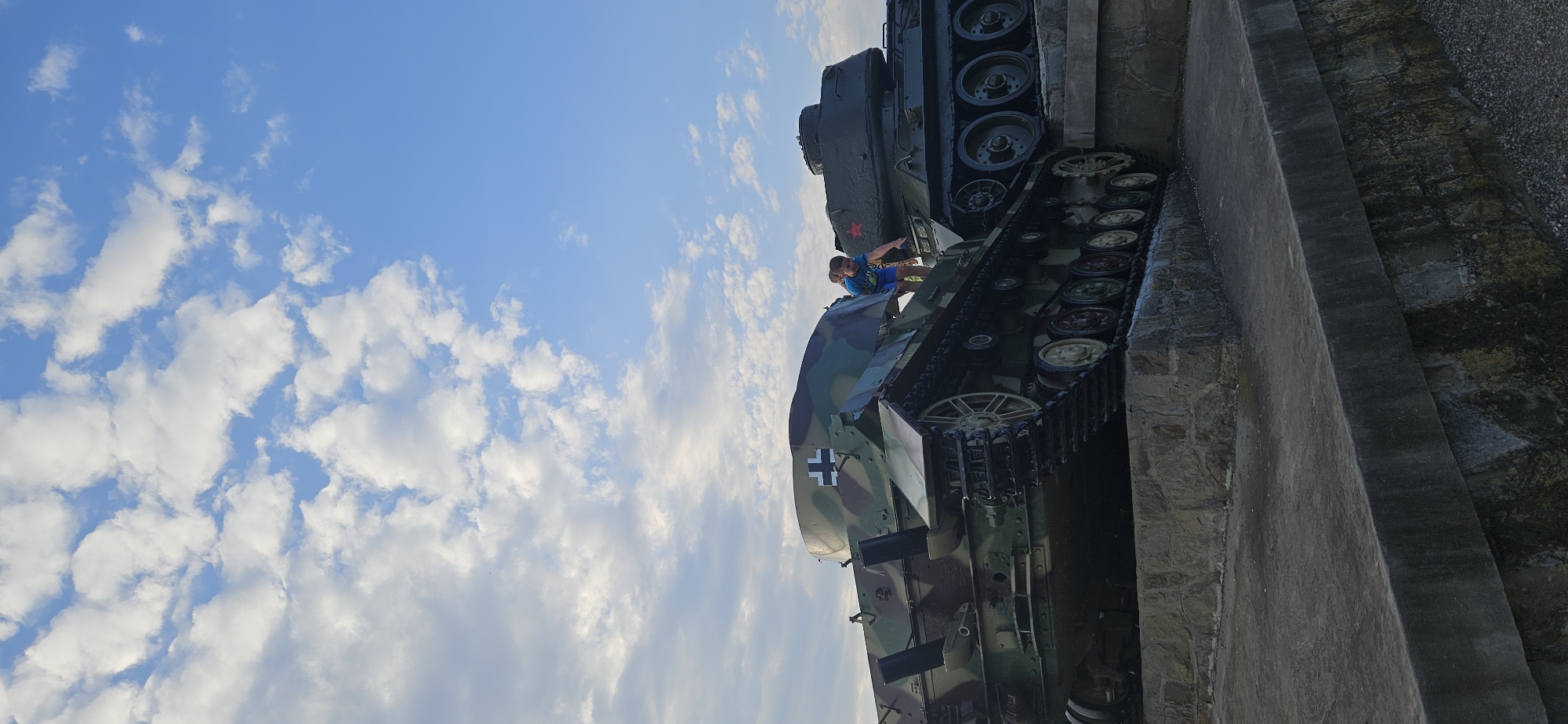 In Svidník, Slovakia, two historical tanks are displayed on a concrete and stone embankment. The tank in the foreground is painted with a camouflage pattern and features a white cross insignia on its turret. A young boy in a blue t-shirt and shorts is climbing on top of this tank, reaching towards the second tank behind it. The second tank is dark green and has a red star insignia on its turret. The sky above is a clear blue with scattered white clouds. The scene appears to be outdoors, possibly in a park or memorial site.