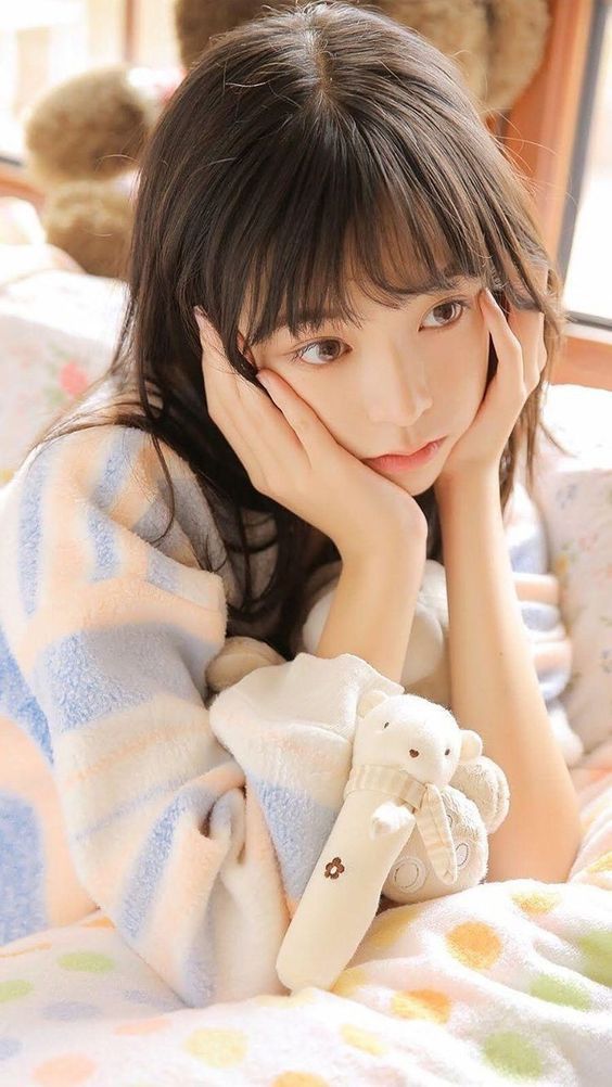 The image shows a young woman with long dark hair sitting on a bed. She is wearing a blue and white striped sweater and is resting her head in her hands, gazing downwards. A stuffed animal is lying next to her. The bed is covered in a patterned blanket. The coordinates provided, -7.45472, 111.427584, locate a point in the city of Geneng, Indonesia. The scene is likely a bedroom in a private residence.