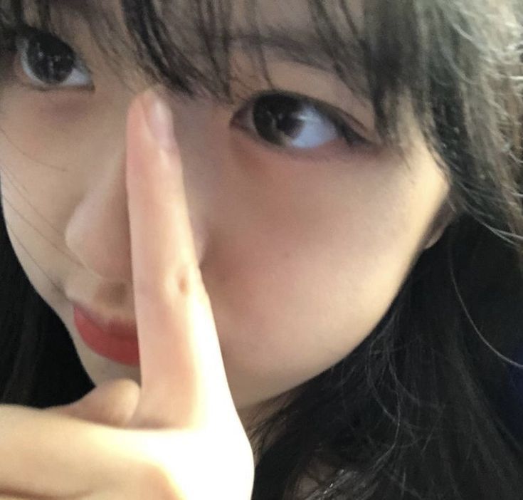 The image shows a close-up of a young woman's face. She has long, dark hair that is parted in the middle and covers part of her forehead. Her right eye is visible, looking directly at the camera. Her right hand is in front of her face, with her index finger covering her left eye and her thumb touching her cheek. Her lips are painted with red lipstick. The coordinates provided in the prompt place the image in Geneng, Indonesia, but there are no specific details about the environment that would confirm this location. It's possible the image was taken in a different location, or the information about the location may be inaccurate.
