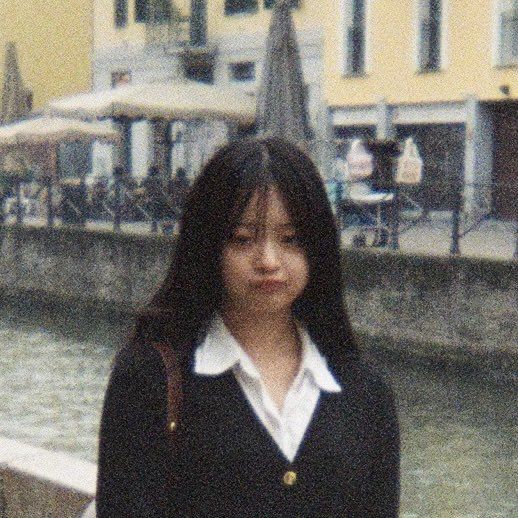 The image shows a young woman with long black hair, wearing a black blazer over a white collared shirt, standing in front of a canal. The woman is looking directly at the camera with a slightly surprised expression. She is wearing a brown leather shoulder bag. The background is blurred and shows a canal with a stone wall on either side. There are buildings in the distance, some with windows and others with a yellow façade. The location is unclear. The image appears to have been taken with a camera phone and the quality is somewhat grainy.