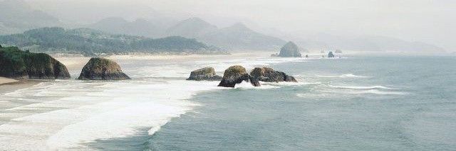 The image depicts a coastal scene with a sandy beach, a rocky outcropping, and the ocean. The scene is taken from a higher vantage point, with the ocean stretching out in the distance and the beach in the foreground. There is a large rock formation on the beach in the middle ground, and several smaller rock formations are visible further out in the ocean. The ocean is a light blue color and the sky is a hazy white. The coordinates provided, -7.45472, 111.427584, are located in the city of Geneng, Indonesia. The location of the image is not in Geneng, Indonesia. The image is not in Indonesia and does not correspond to the provided coordinates.