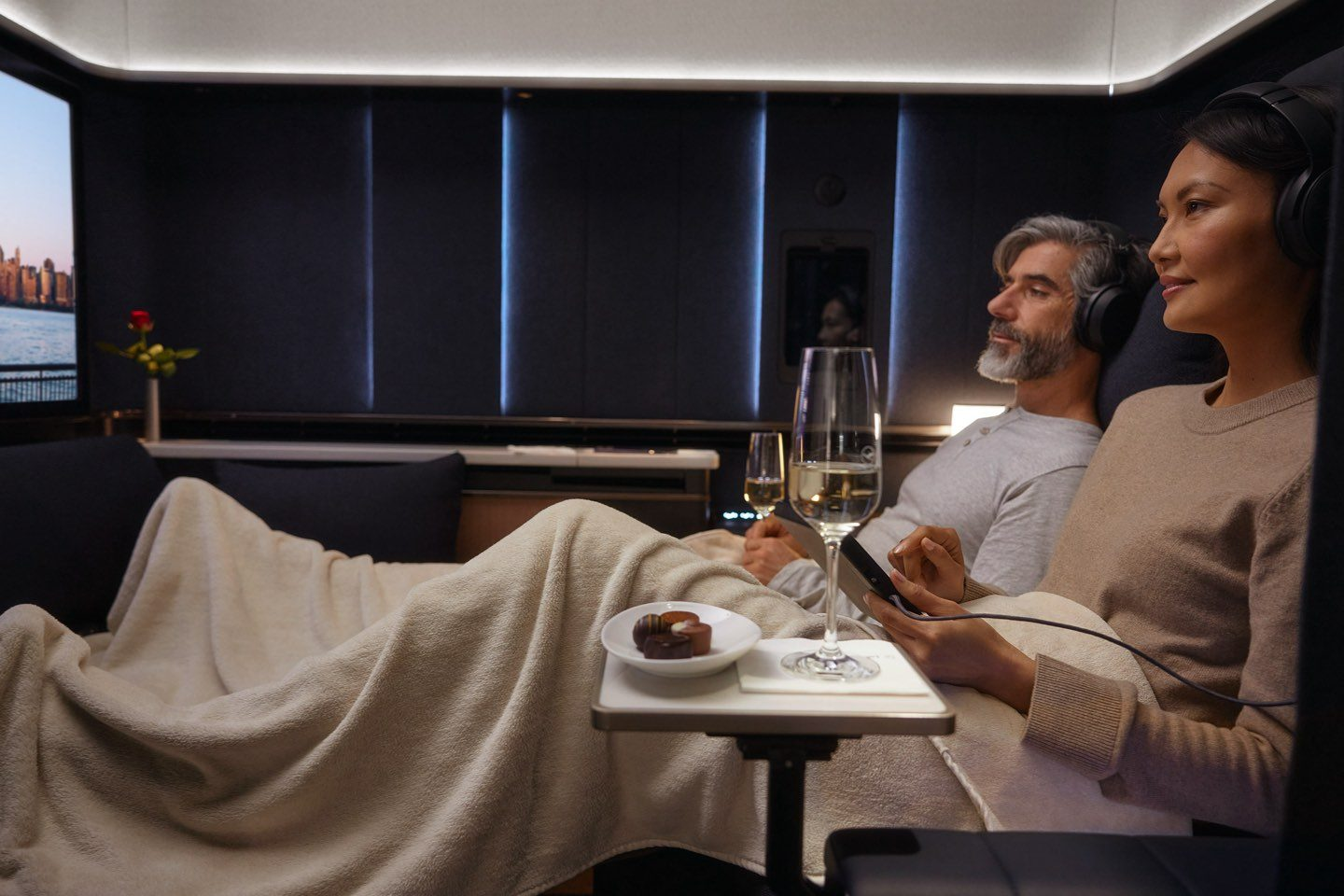 Zwei Passagiere sitzen dicht an dicht in der First-Class-Suite der Lufthansa, ein Weinglas balanciert wackelig auf einem schmalen Beistelltisch.