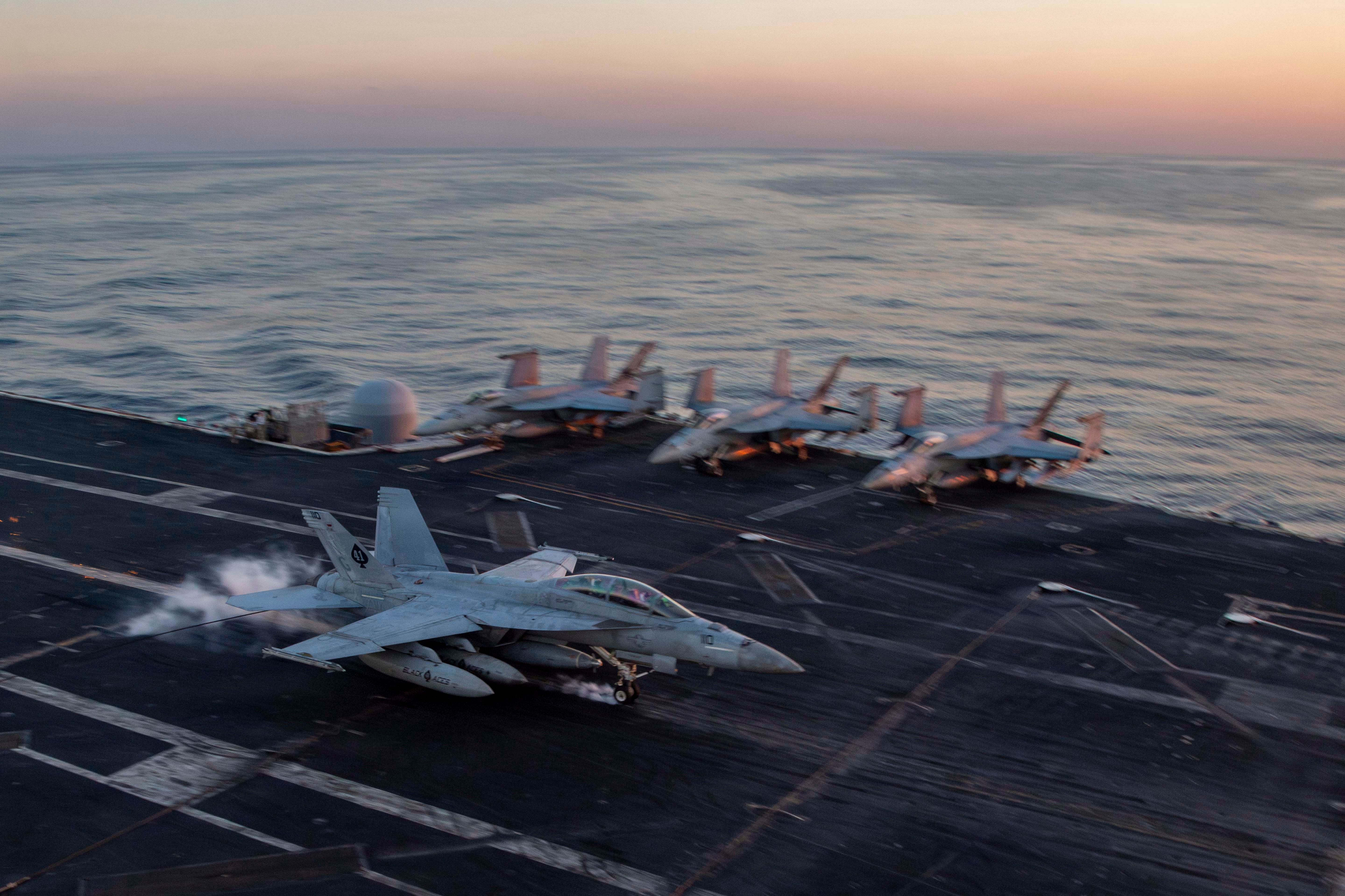 F/A-18E Super Hornet landing on a U.S. Navy aircraft carrier deck at sunset.