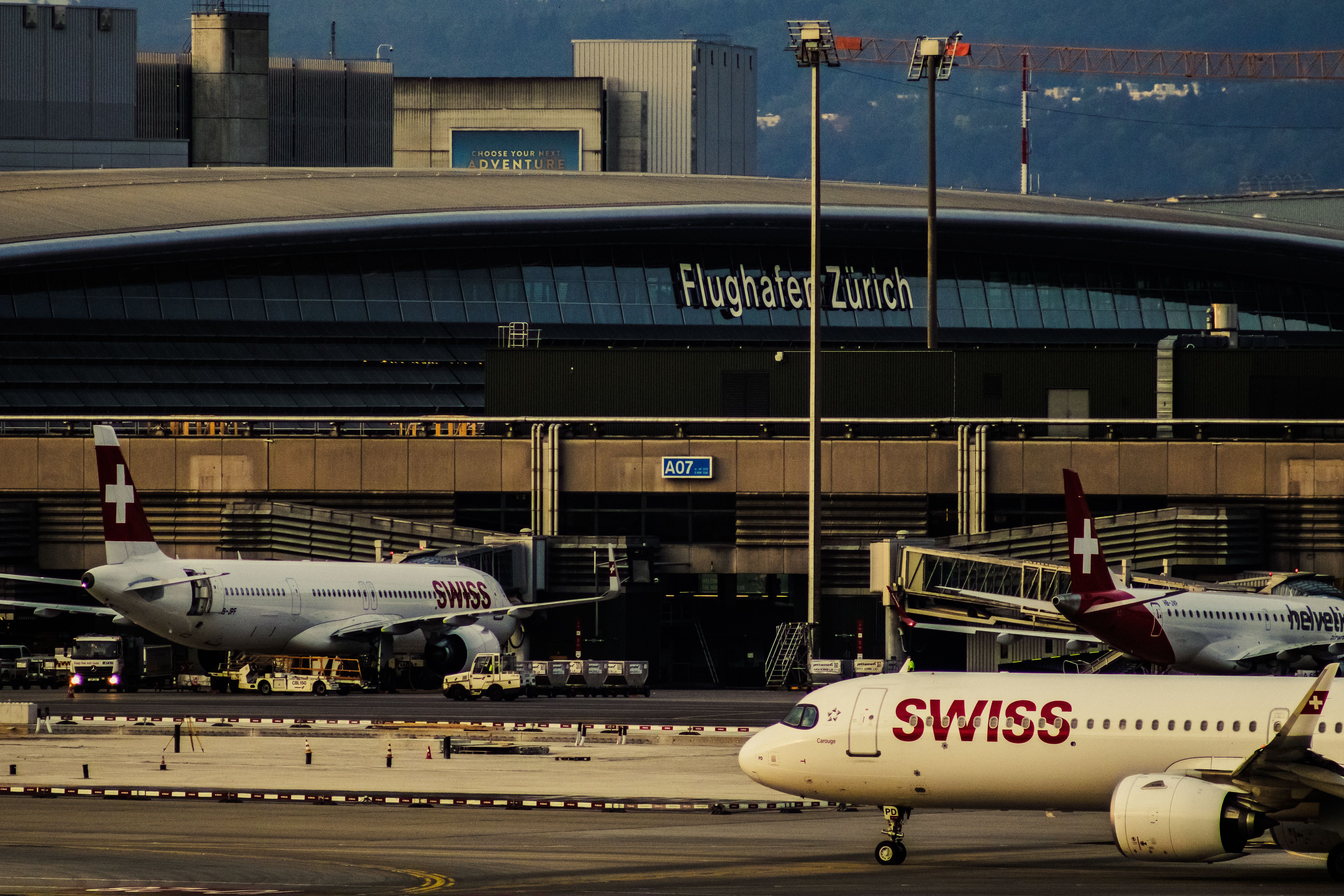 Two SWISS aircraft, including 'Carouge', on the tarmac at Flughafen Zürich, with a "CHOOSE YOUR NEXT ADVENTURE" sign on the terminal.