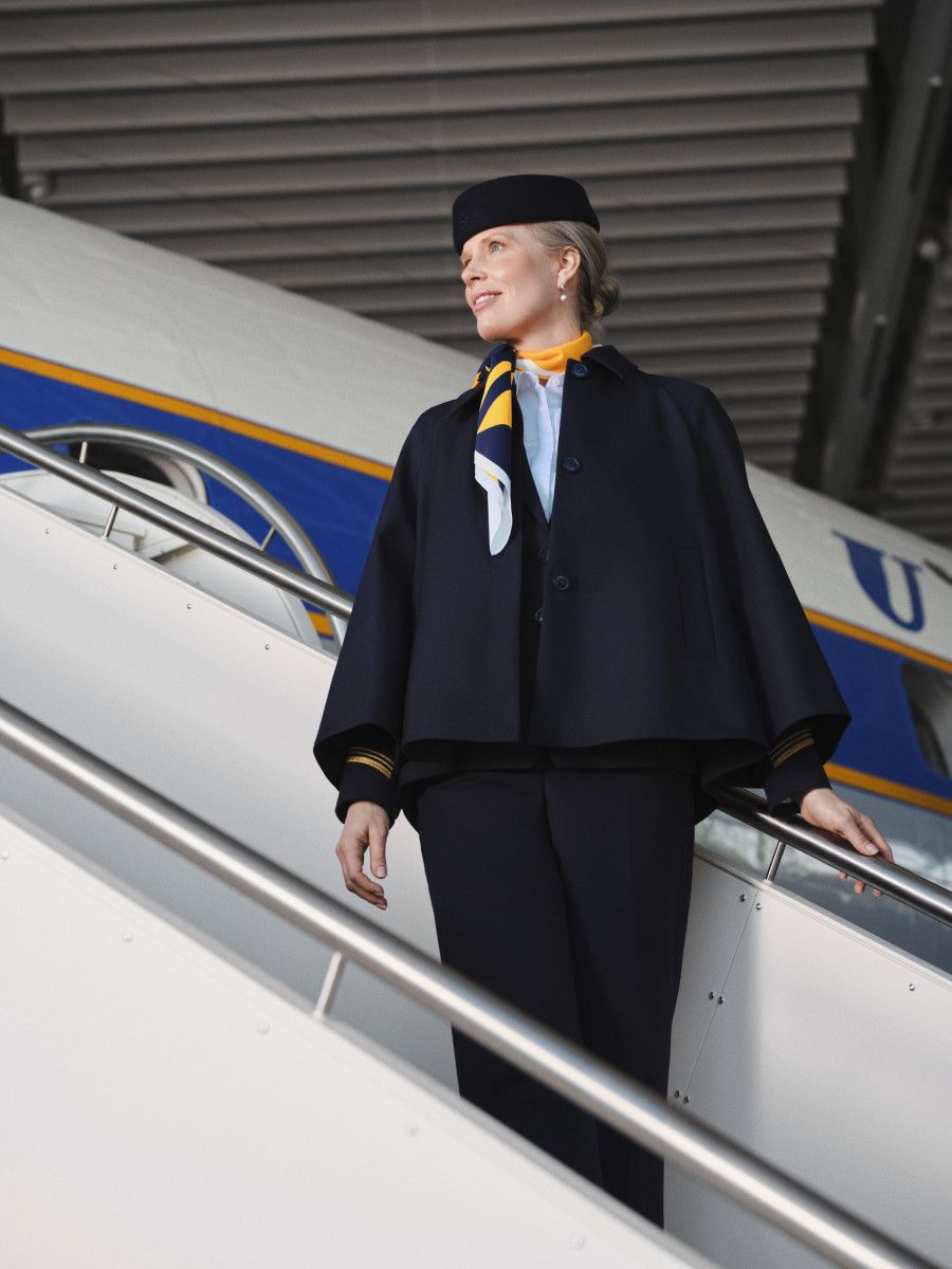 Eine Flugbegleiterin in dunkelblauer Cape-Uniform steht auf einer Flugzeugtreppe und blickt zur Seite.