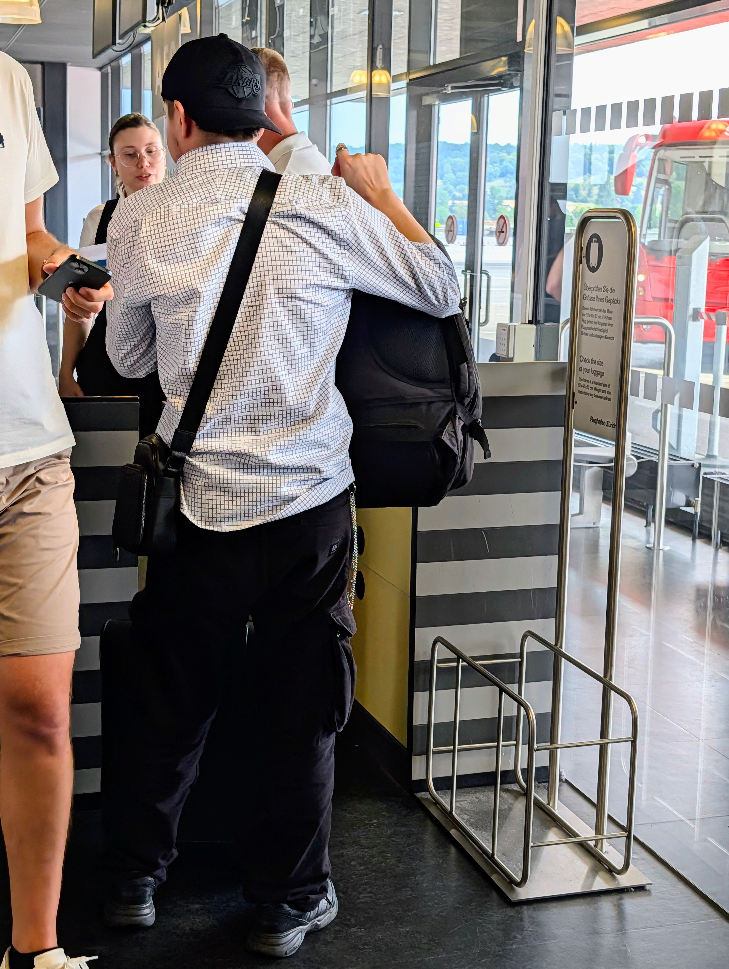 Reisende überprüfen Gepäckgröße am Flughafen Zürich. Eine Person mit Lakers-Kappe spricht mit Personal neben einer Gepäckschablone.