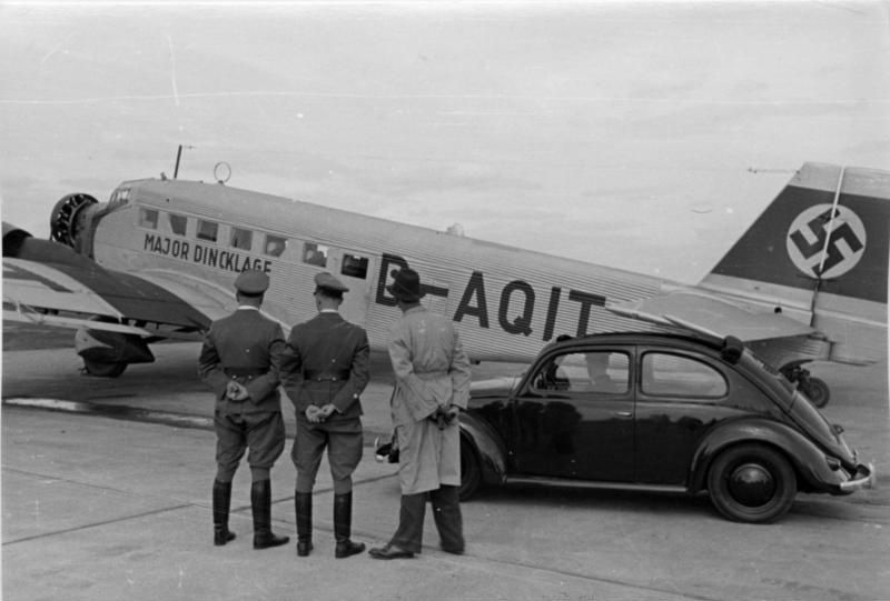 Drei Männer stehen vor einem Junkers Ju 52 Flugzeug und einem VW Käfer auf einem Flugplatz in der Zeit des Nationalsozialismus.