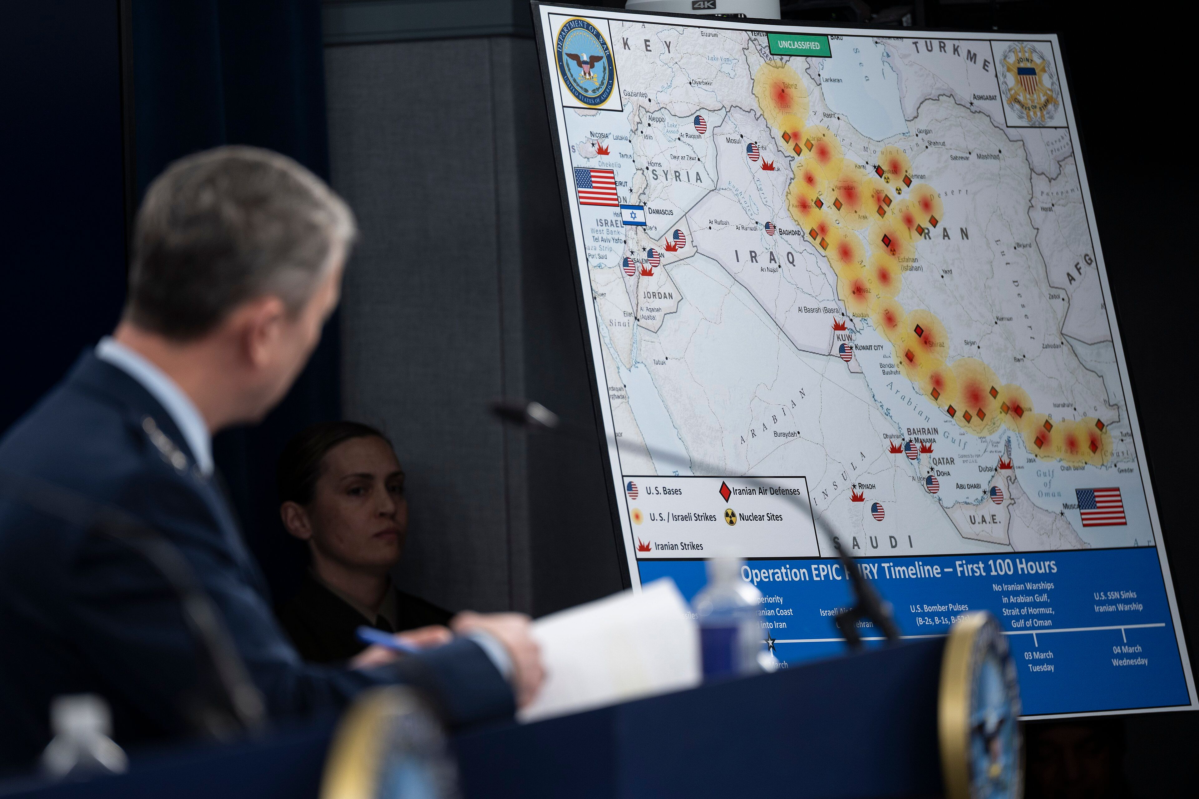 Briefing-Karte mit Angriffen im Nahen Osten mit US- und iranischen Flaggen, Beamte im Vordergrund.